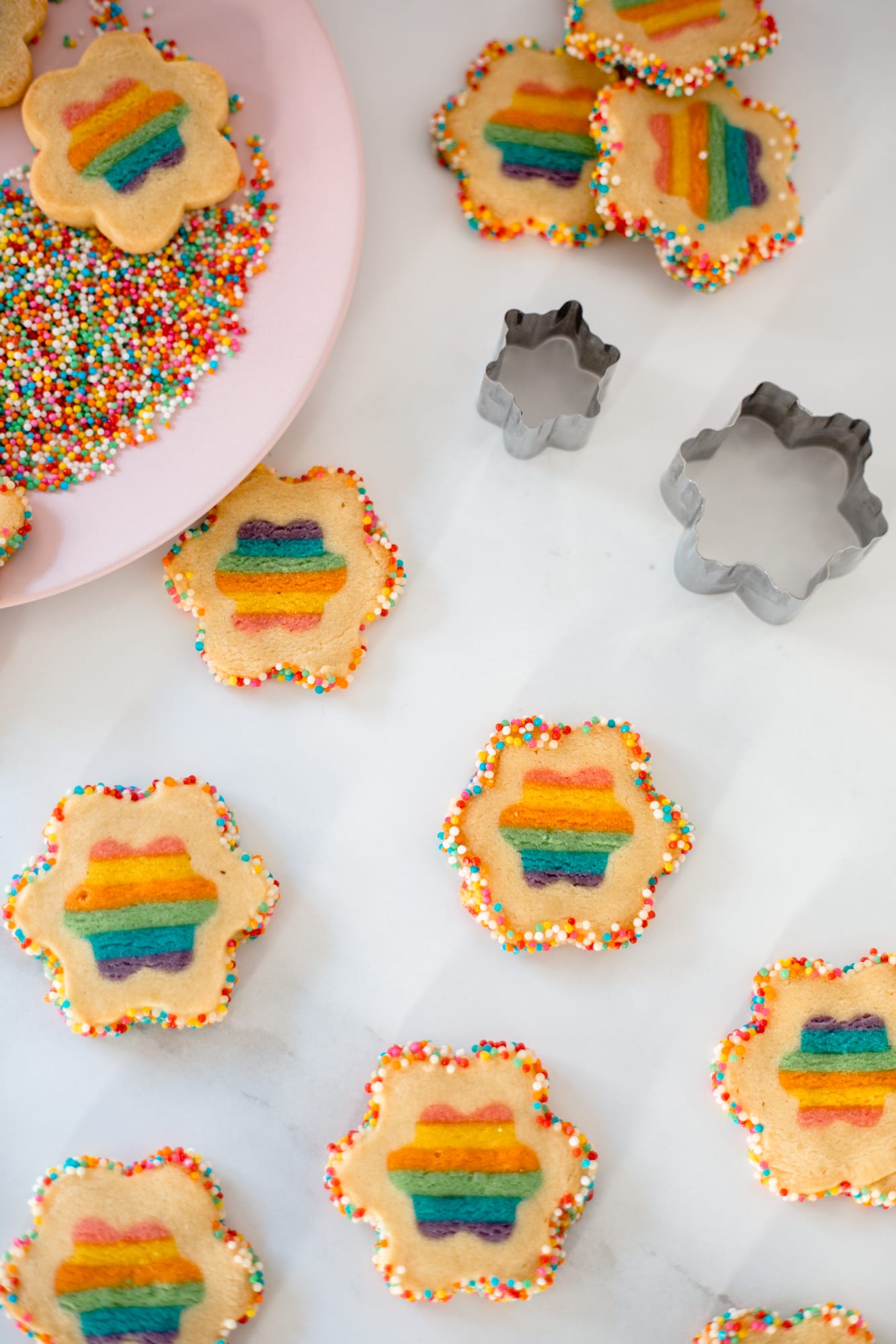 Rainbow cookies beside a pink plate of sprinkles and two flower cutters