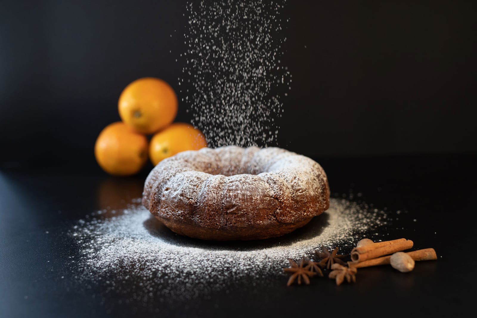 Icing sugar falling over an orange spice pound cake baked in a bundt tin, with oranges and spices around it
