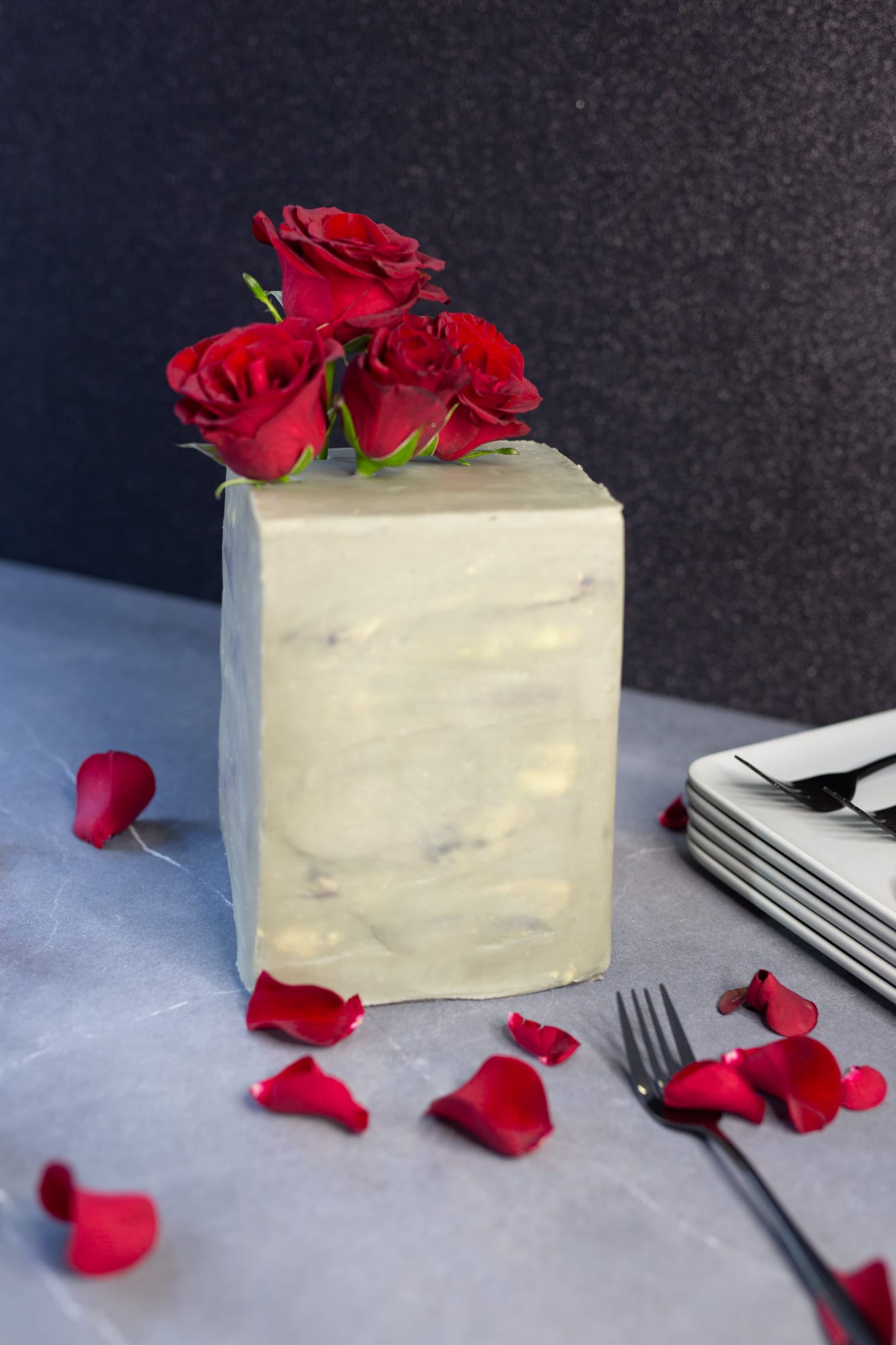 A tall decadent red velvet cake topped with red roses beside scattered petals, plates and forks
