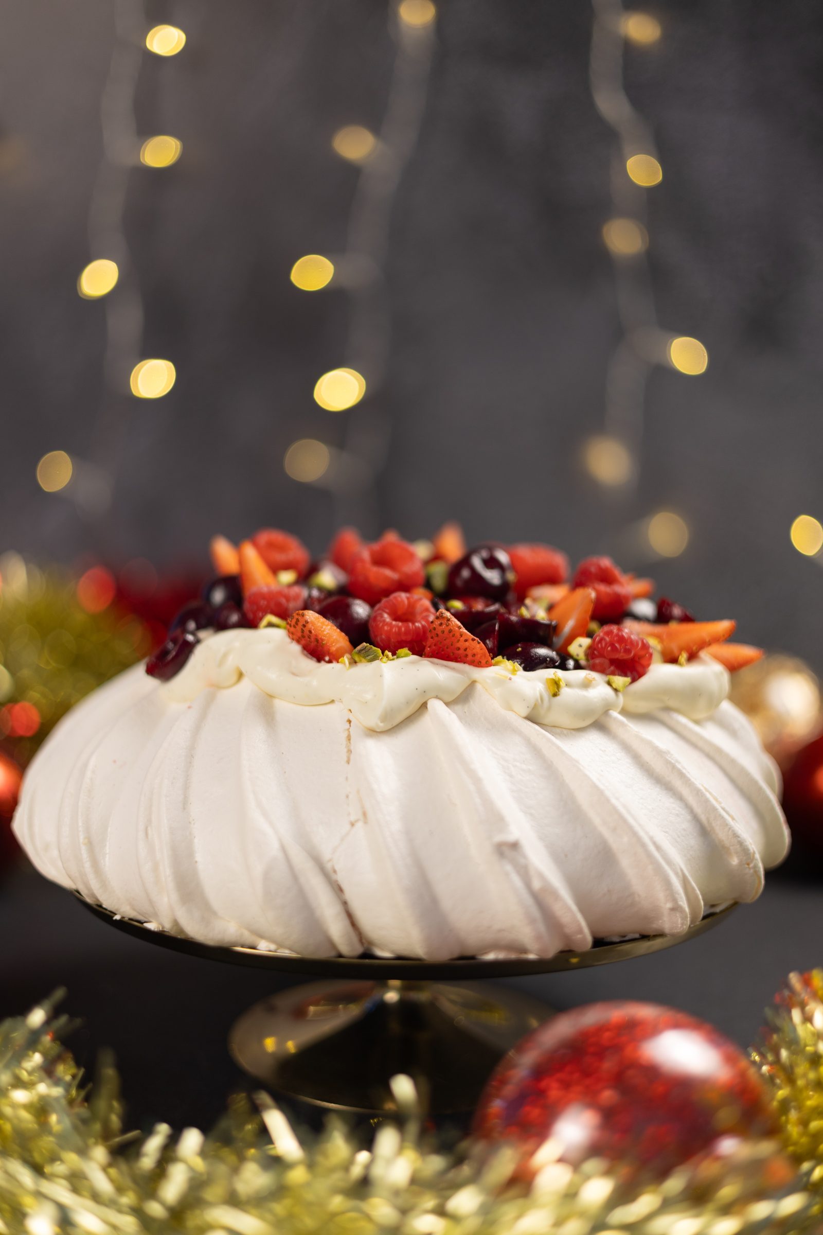 A very berry pavlova on a gold cake stand in front of fairy lights, from the side