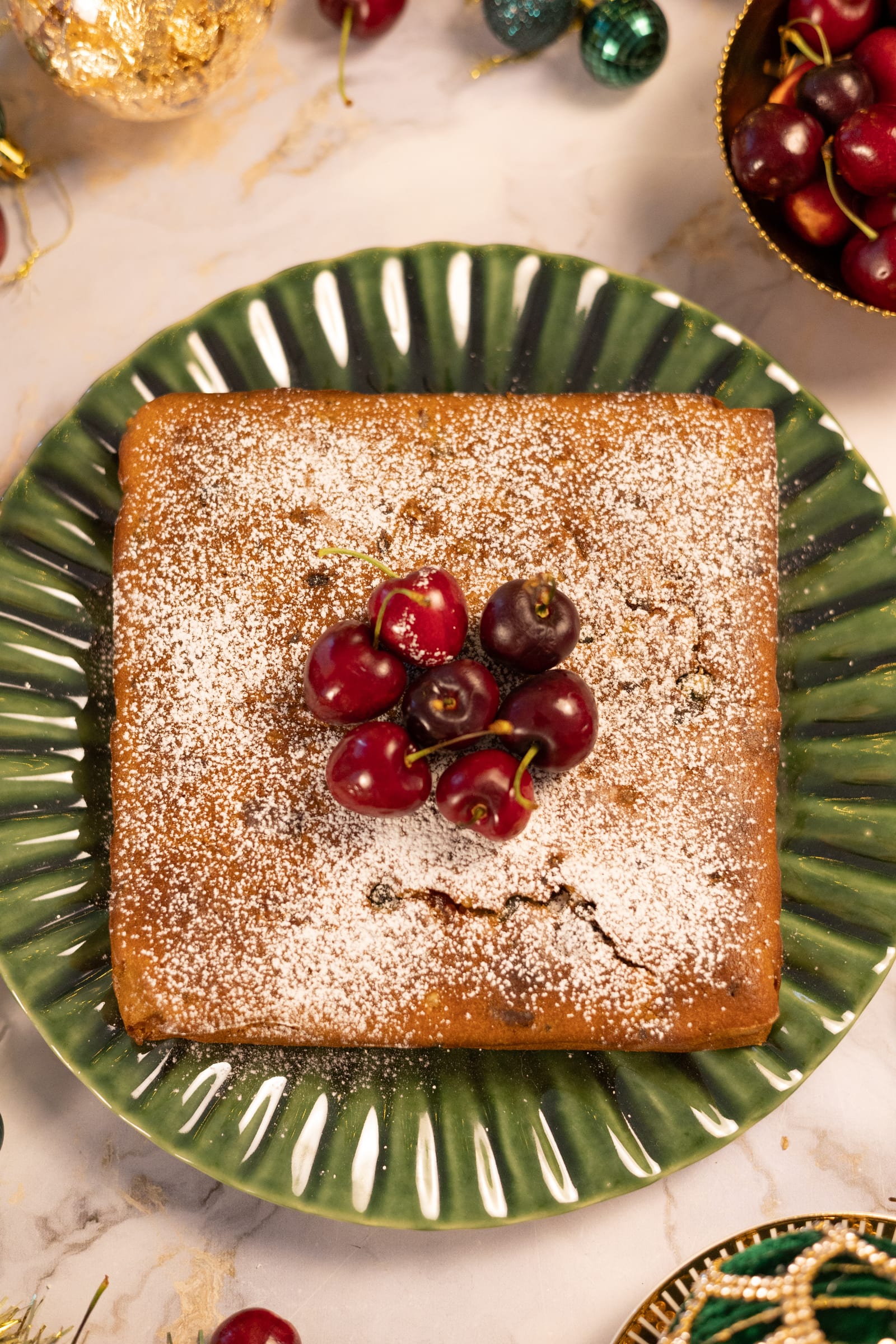 A square fruitcake dusted with icing sugar and topped with cherries