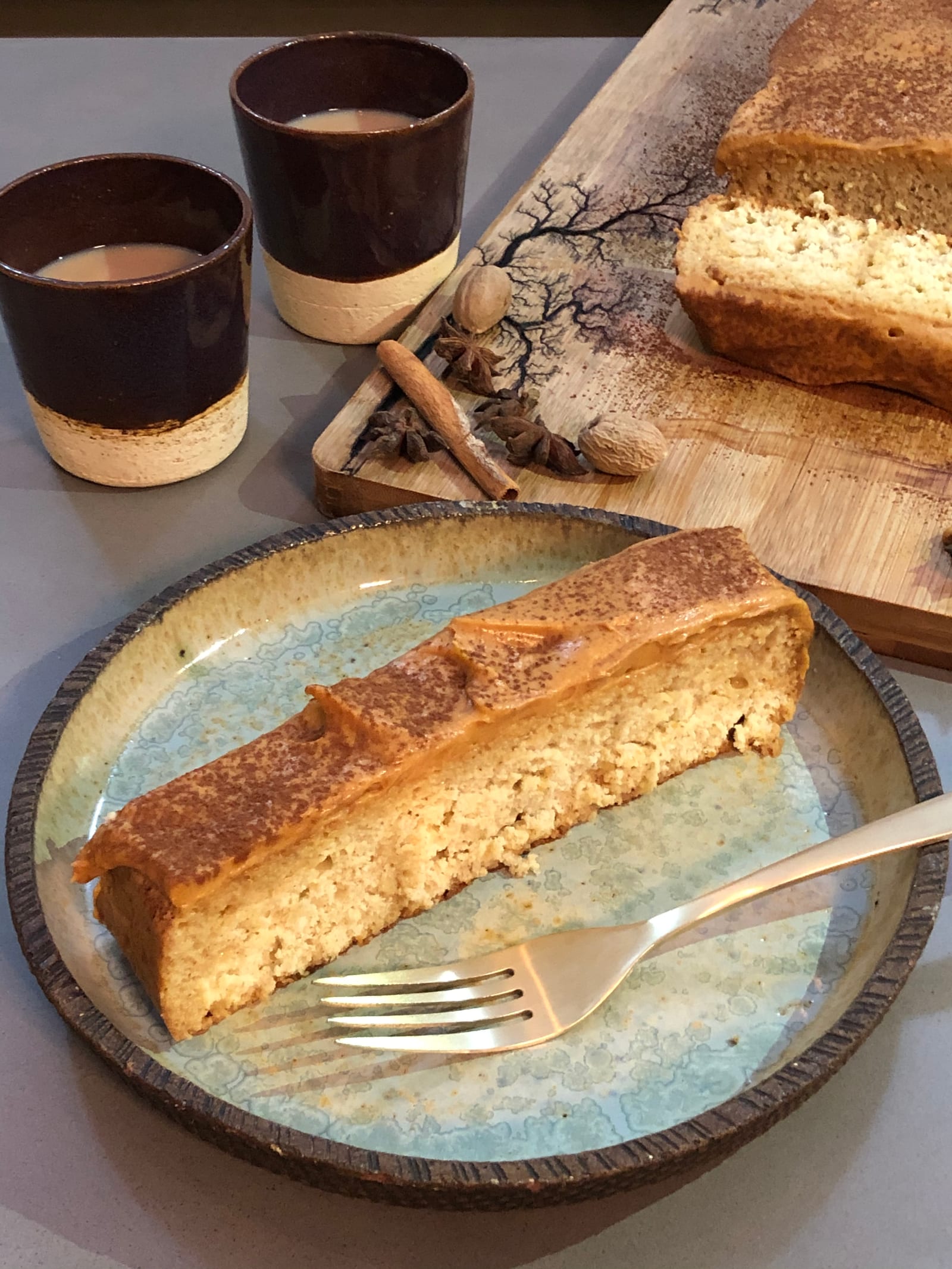 A slice of masala tres leches cake on a blue plate with a fork, cups and the cake behind