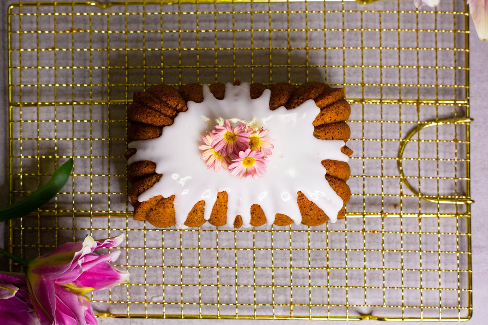 A glazed gluten-free lemon and poppyseed loaf with a pink daisy on a gold wire rack, from above