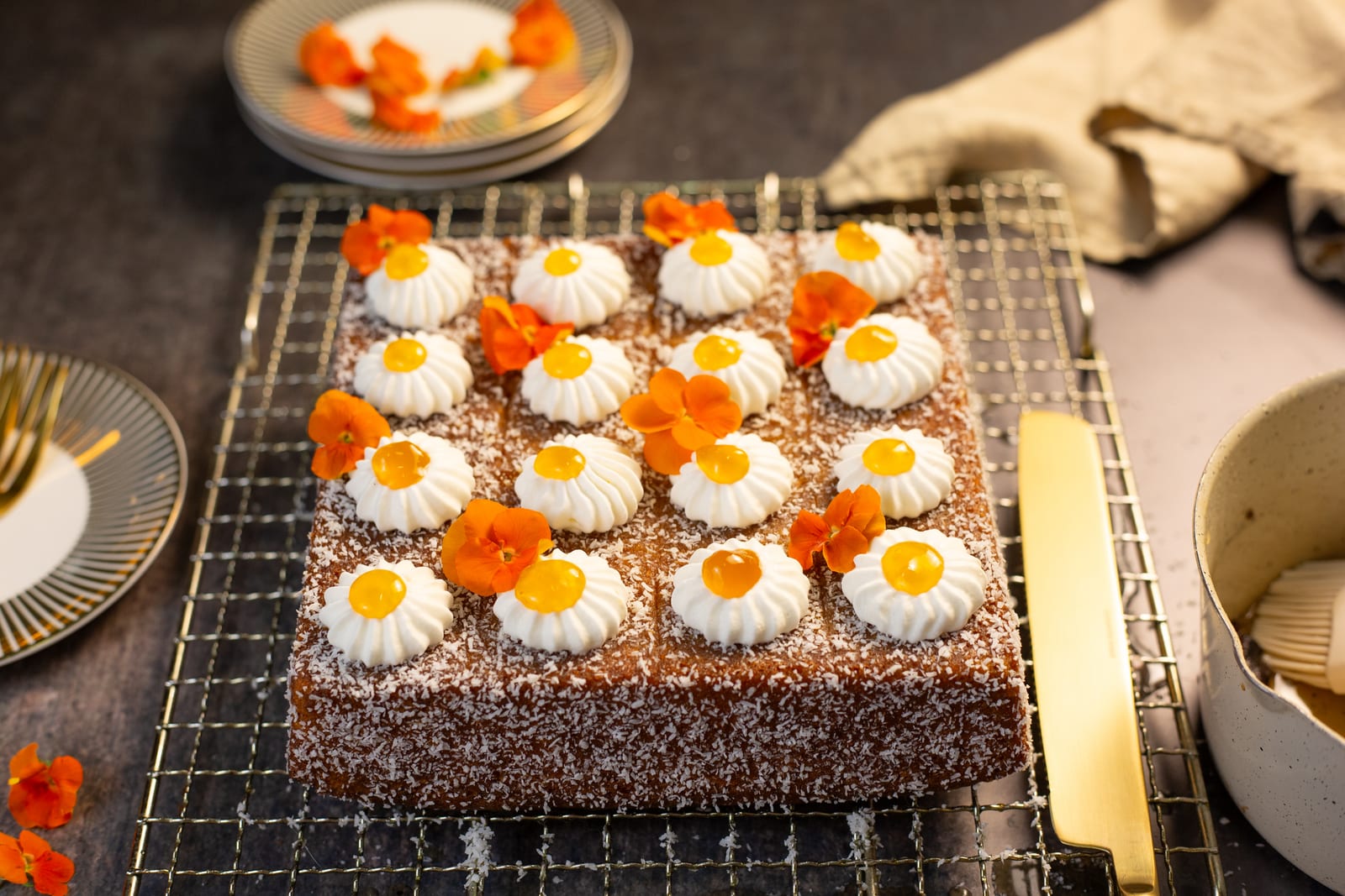 A koesister traybake on a wire rack, topped with rows of cream rosettes and orange flowers