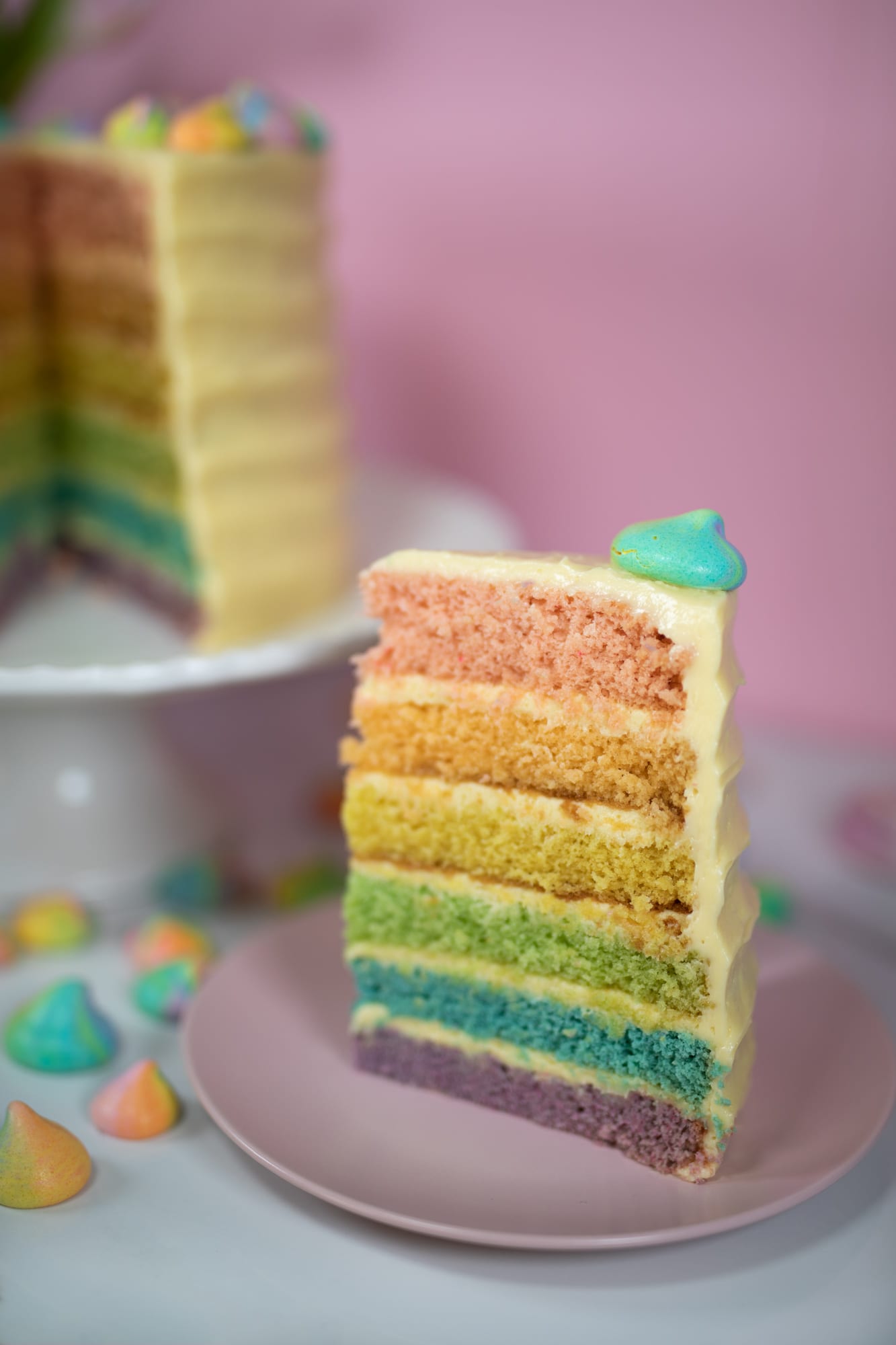 A tall slice of pastel rainbow cake on a pink plate, the cut cake behind