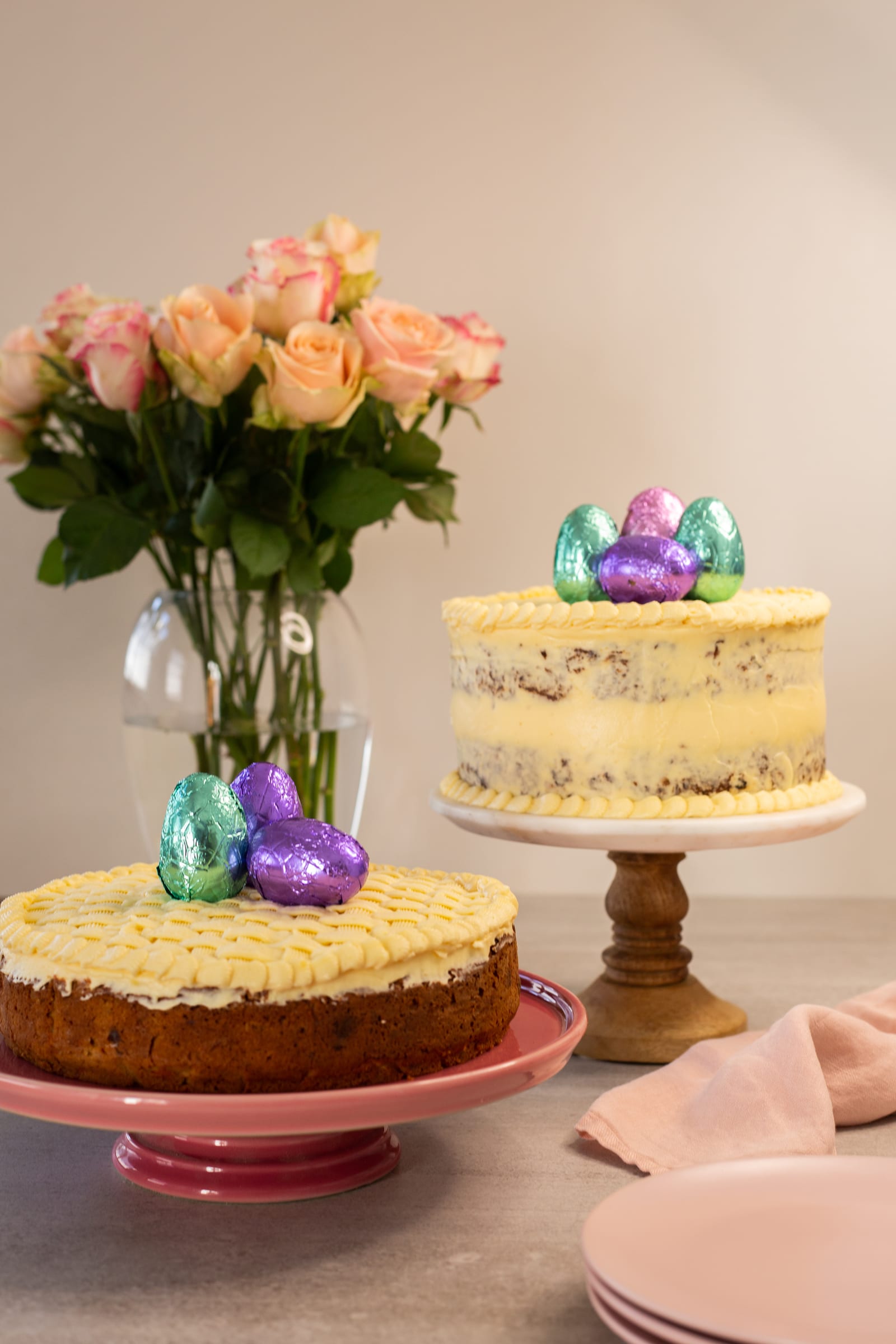 Two gluten-free carrot cakes on pink and wooden stands beside a vase of peach roses