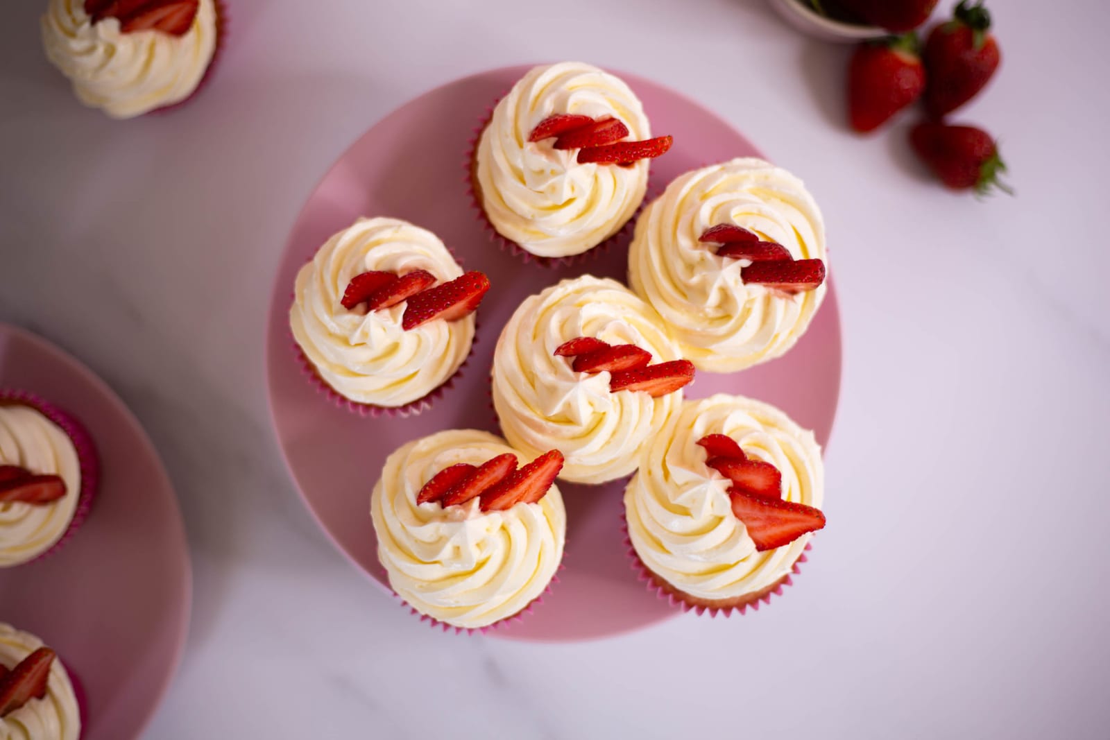 Strawberry and white chocolate cupcakes with swirled buttercream and strawberry halves on a pink plate, from above