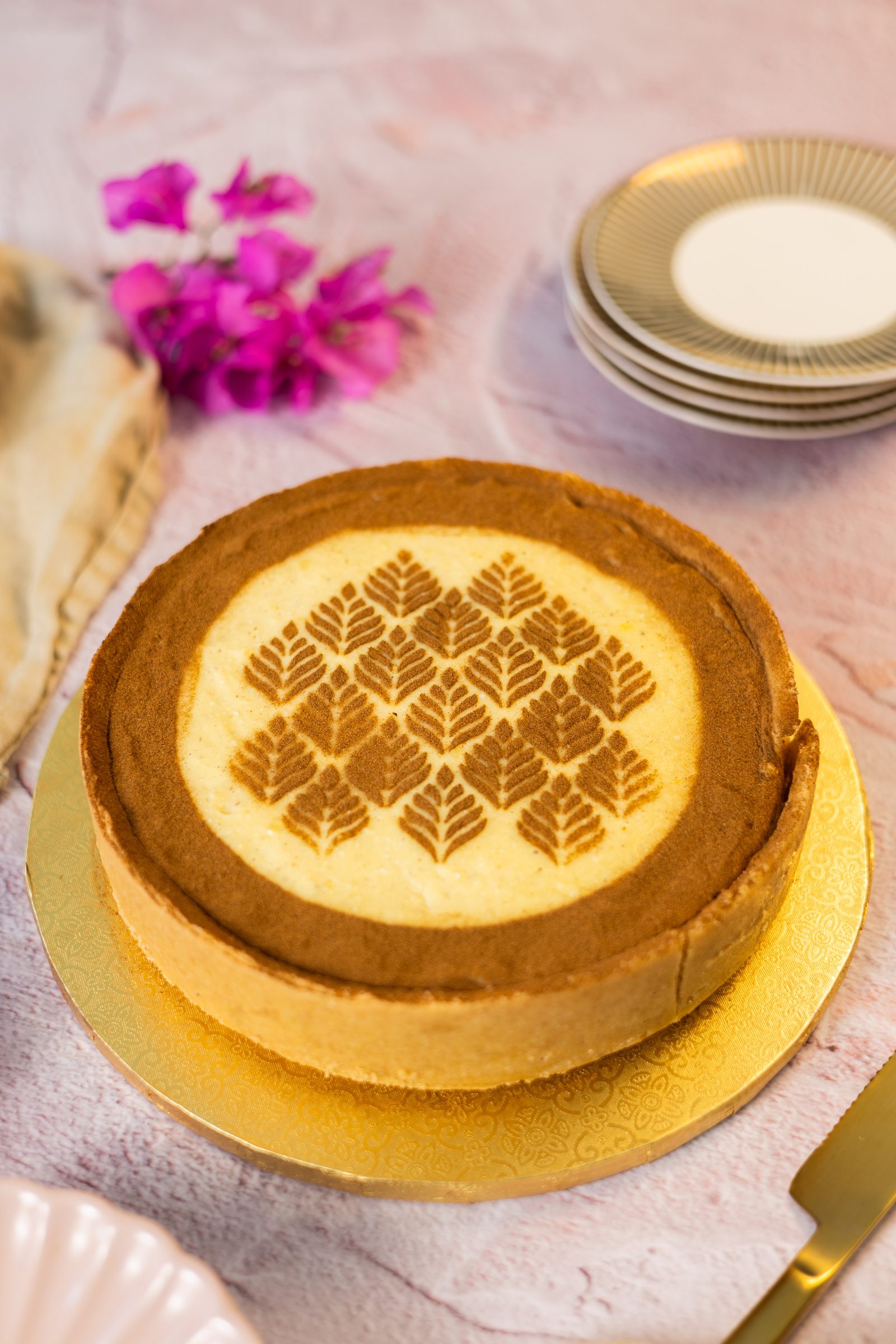 A whole no bake milk tart on a gold cake board beside magenta flowers and a stack of gold-rimmed plates