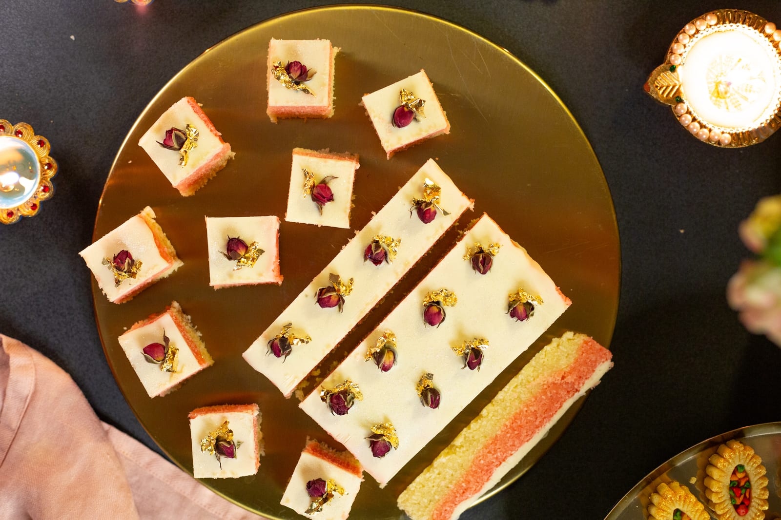 Rose and cardamom coconut kopra paak squares and bars topped with rosebuds on a gold platter beside diyas, from above
