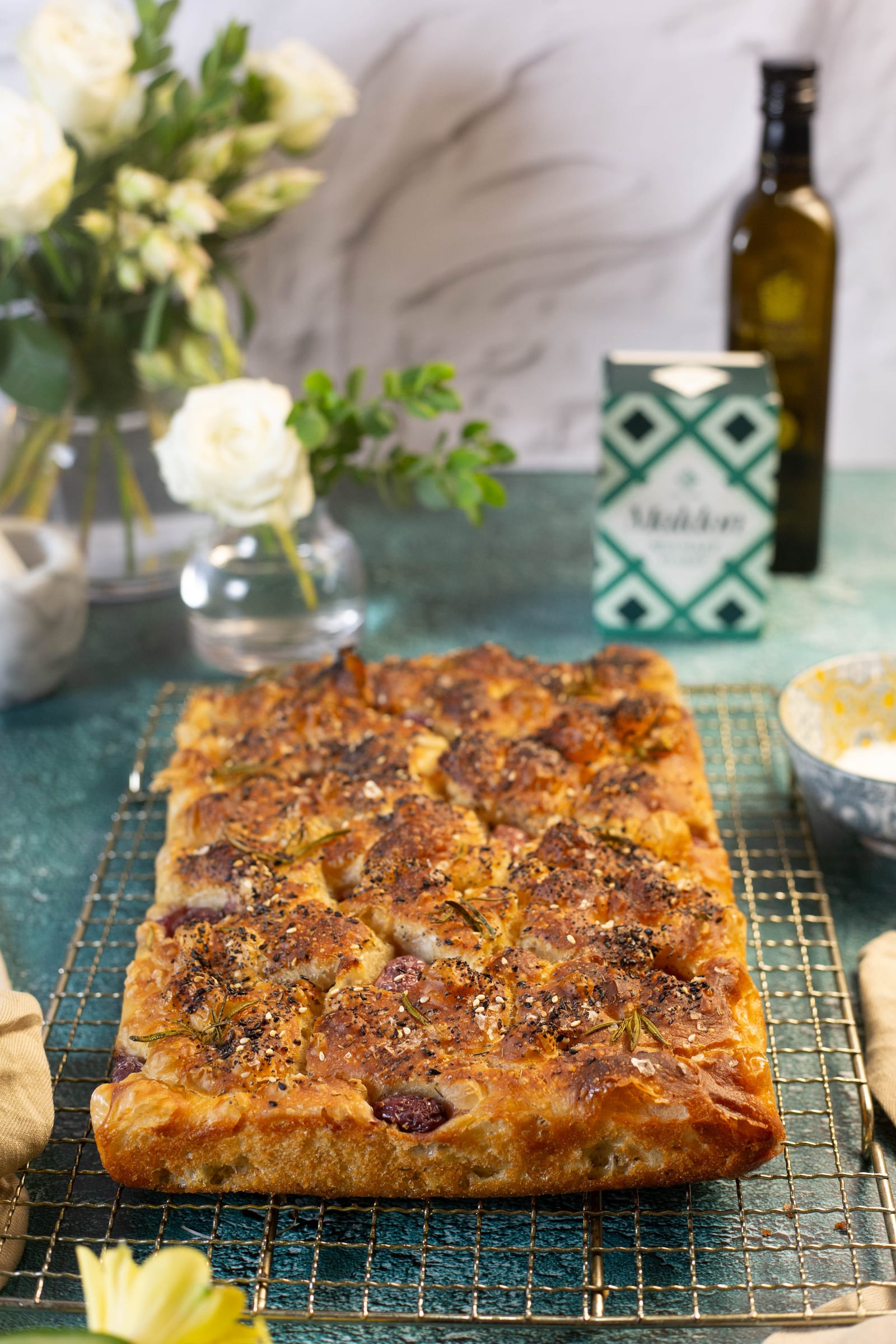 The baked focaccia on a rack, with olive oil and white flowers behind