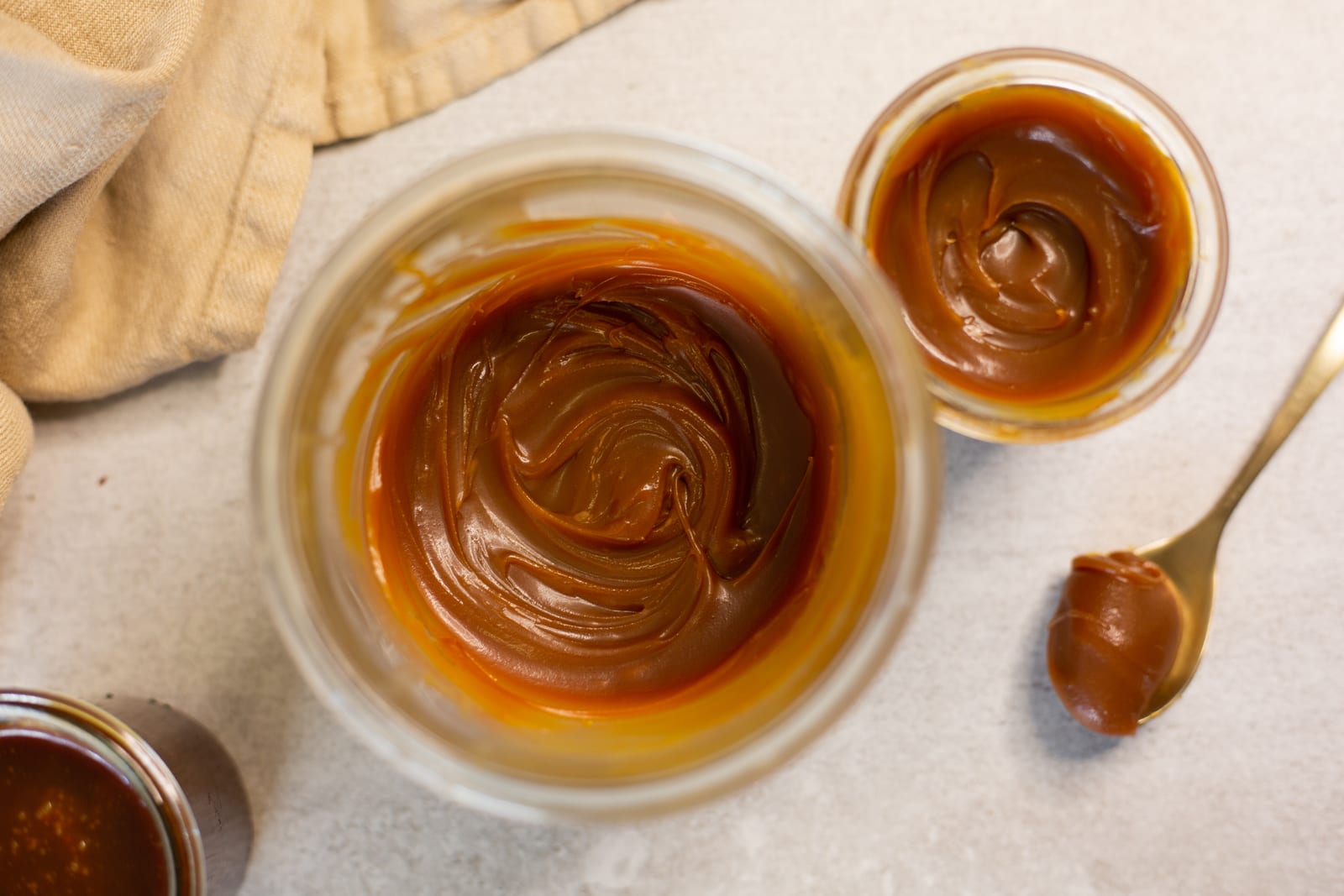 A glass bowl of swirled salted caramel sauce from above, with a small ramekin and a gold spoon