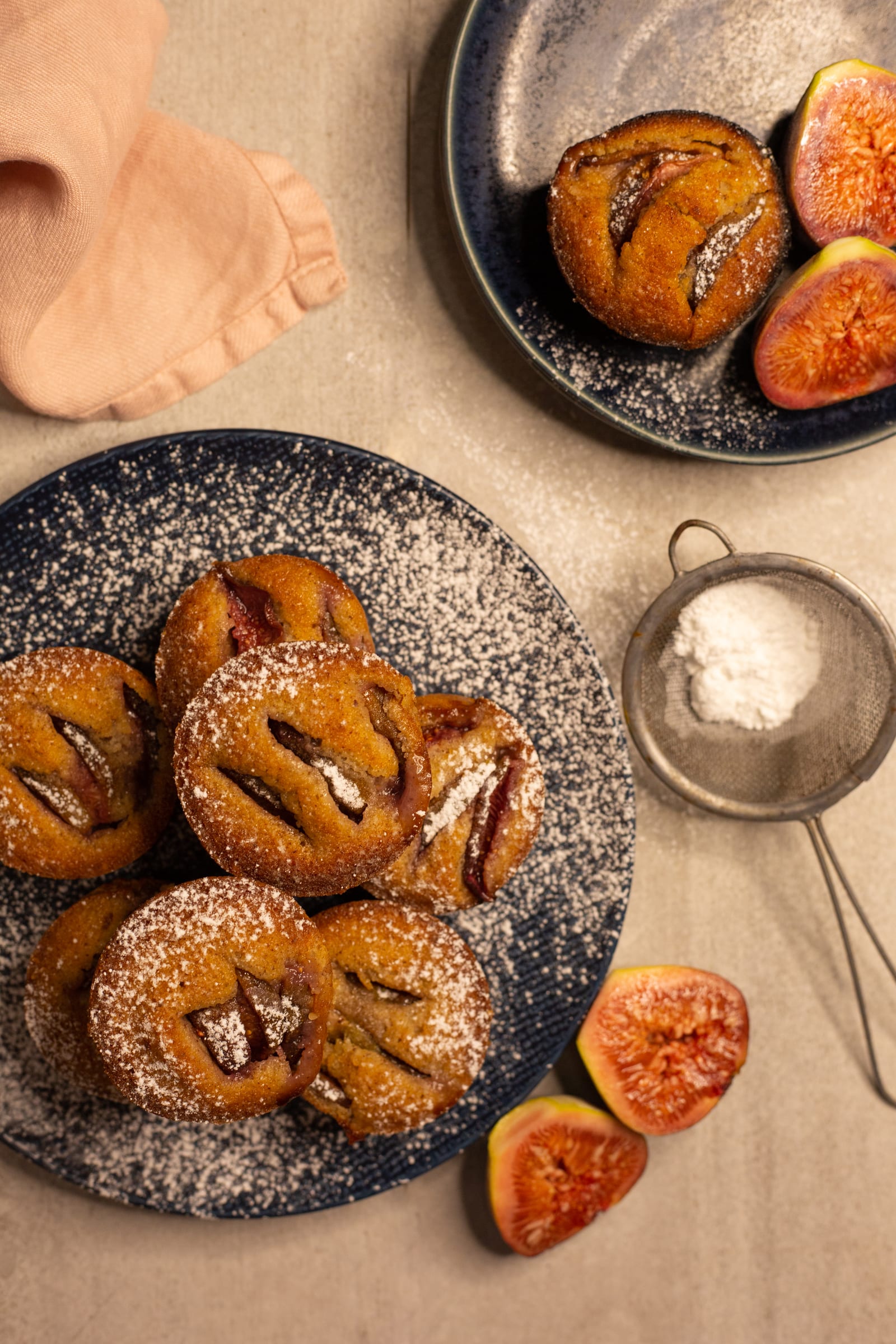 Fig and hazelnut friands on a navy plate beside a sieve and fig halves, from above