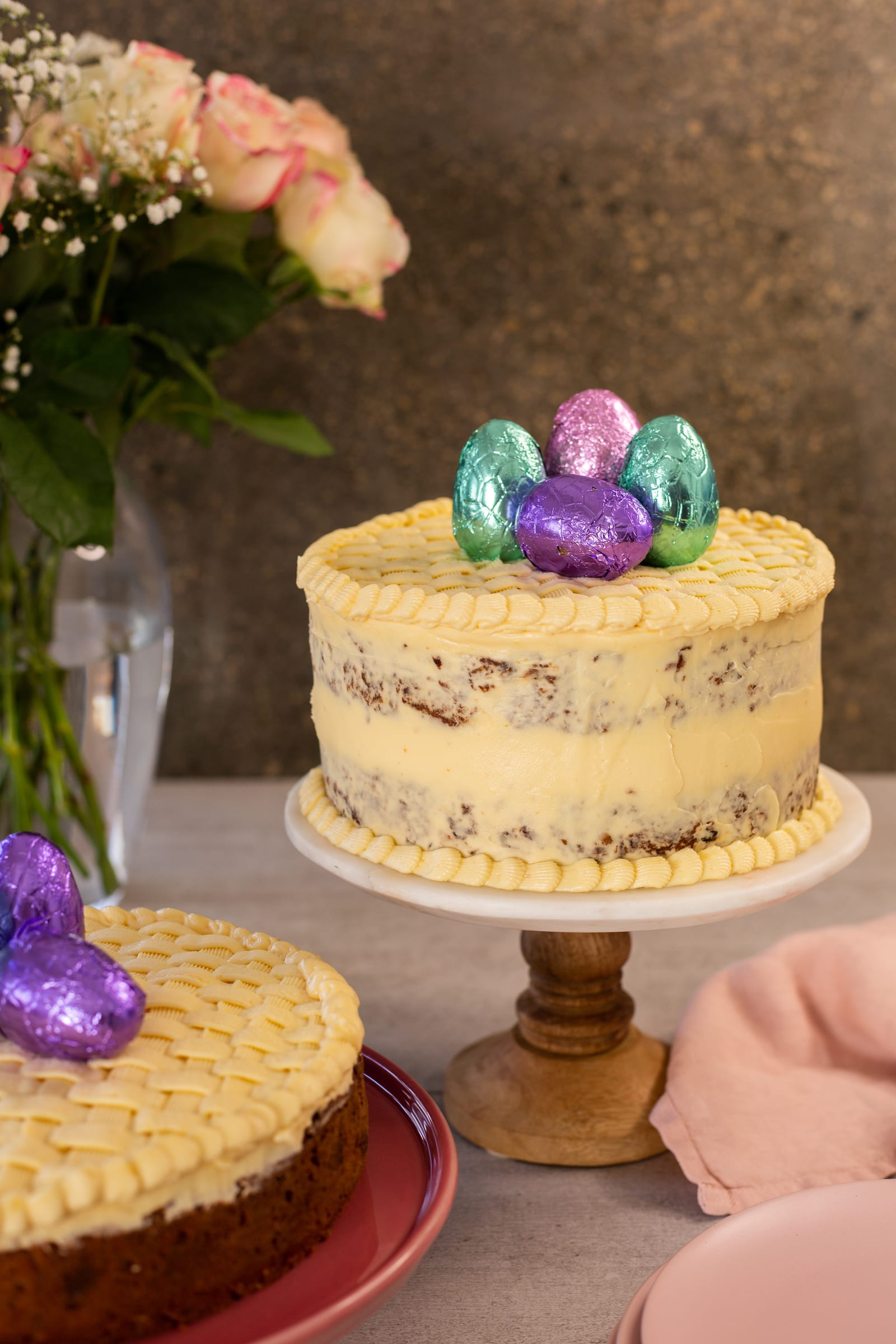 A layered gluten-free carrot cake topped with Easter eggs on a wooden stand, roses behind