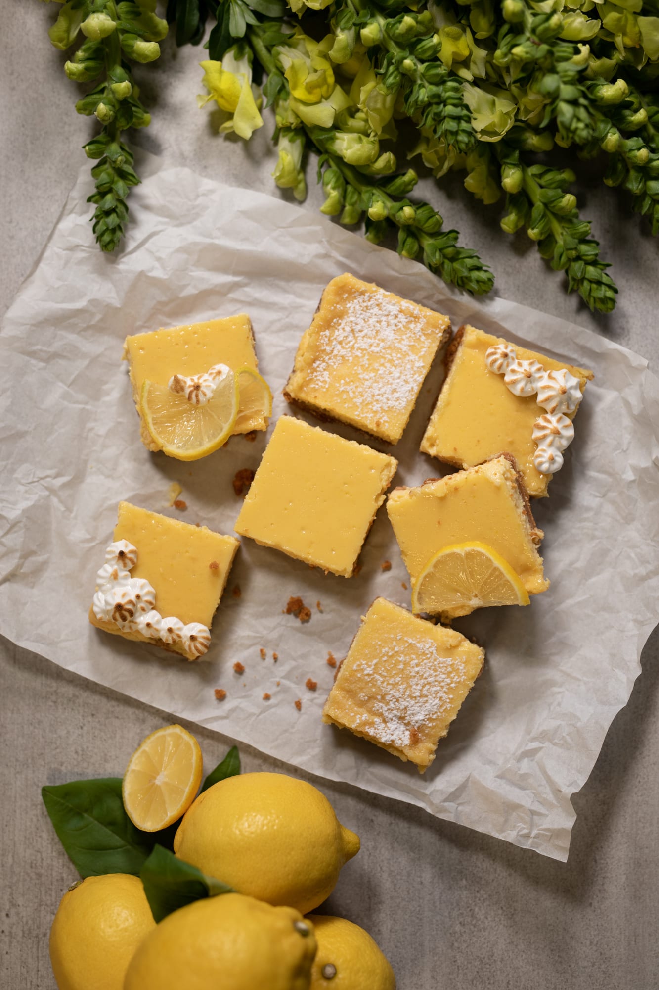 Sweet and tart lemon squares, one topped with a lemon slice, on parchment with lemons, from above