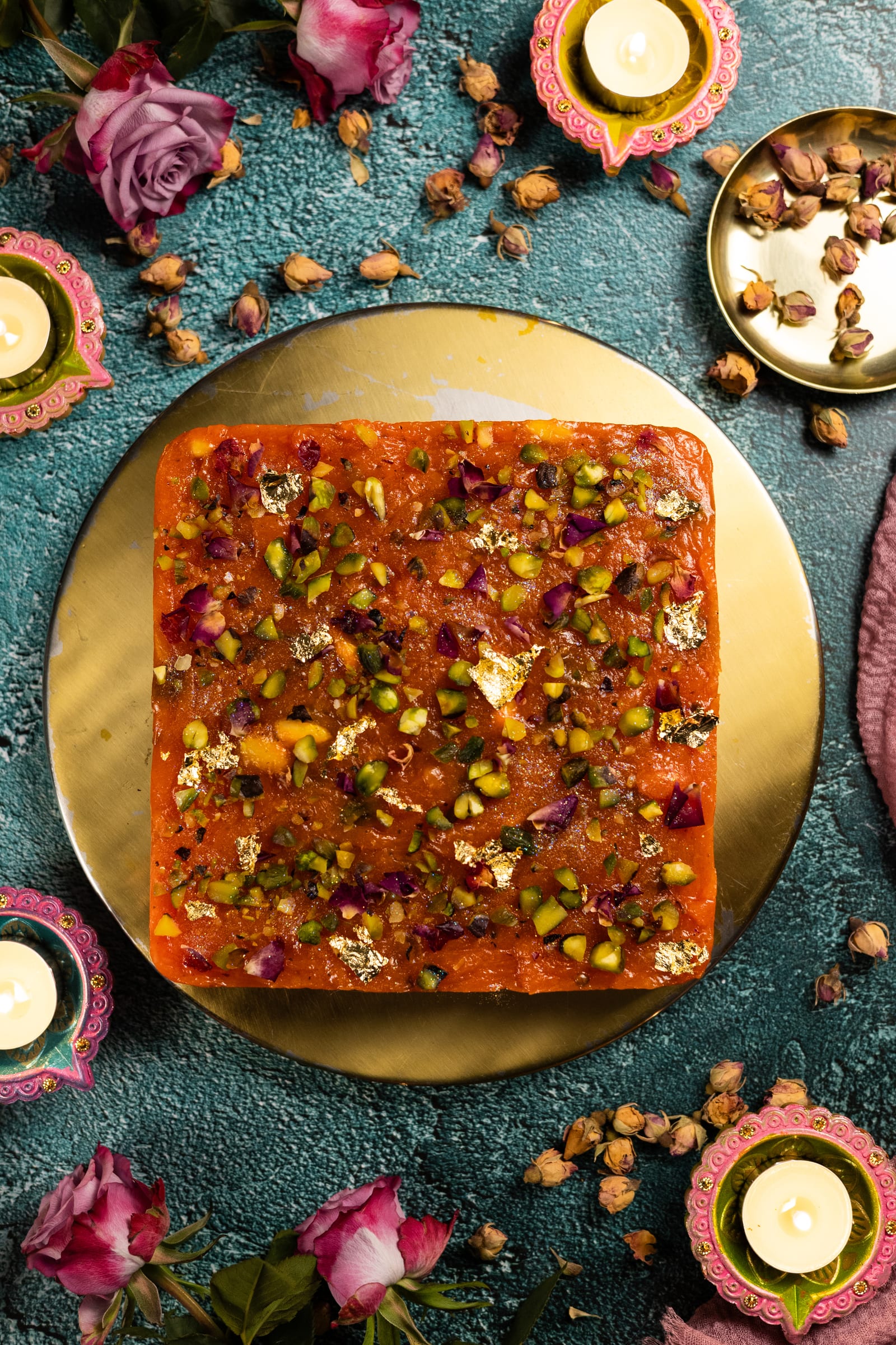An uncut slab of halwa on a gold board with diyas, roses and almonds, from above