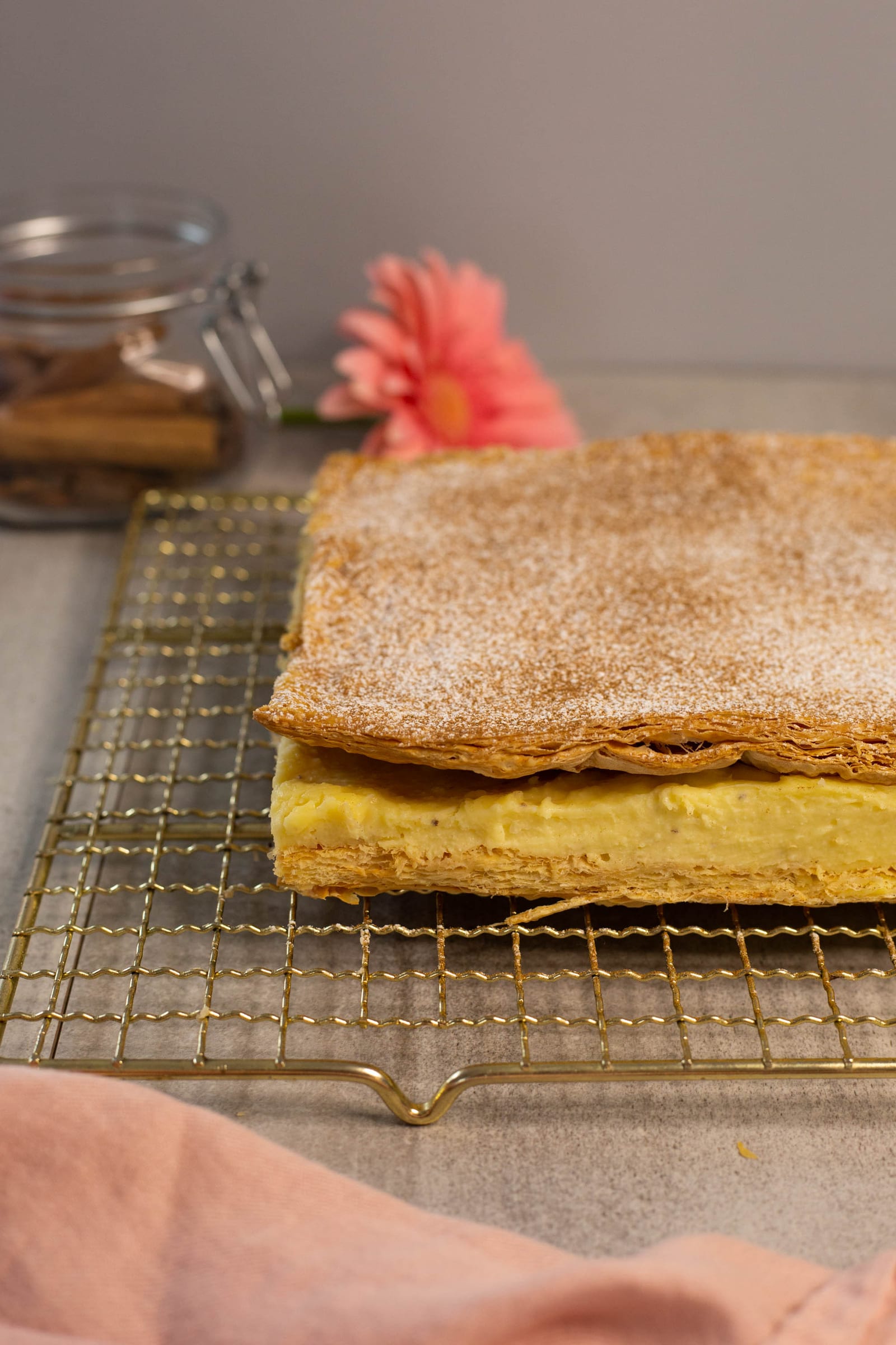 The uncut slab of milk tart slices on a gold wire rack, spice jars and a pink gerbera behind