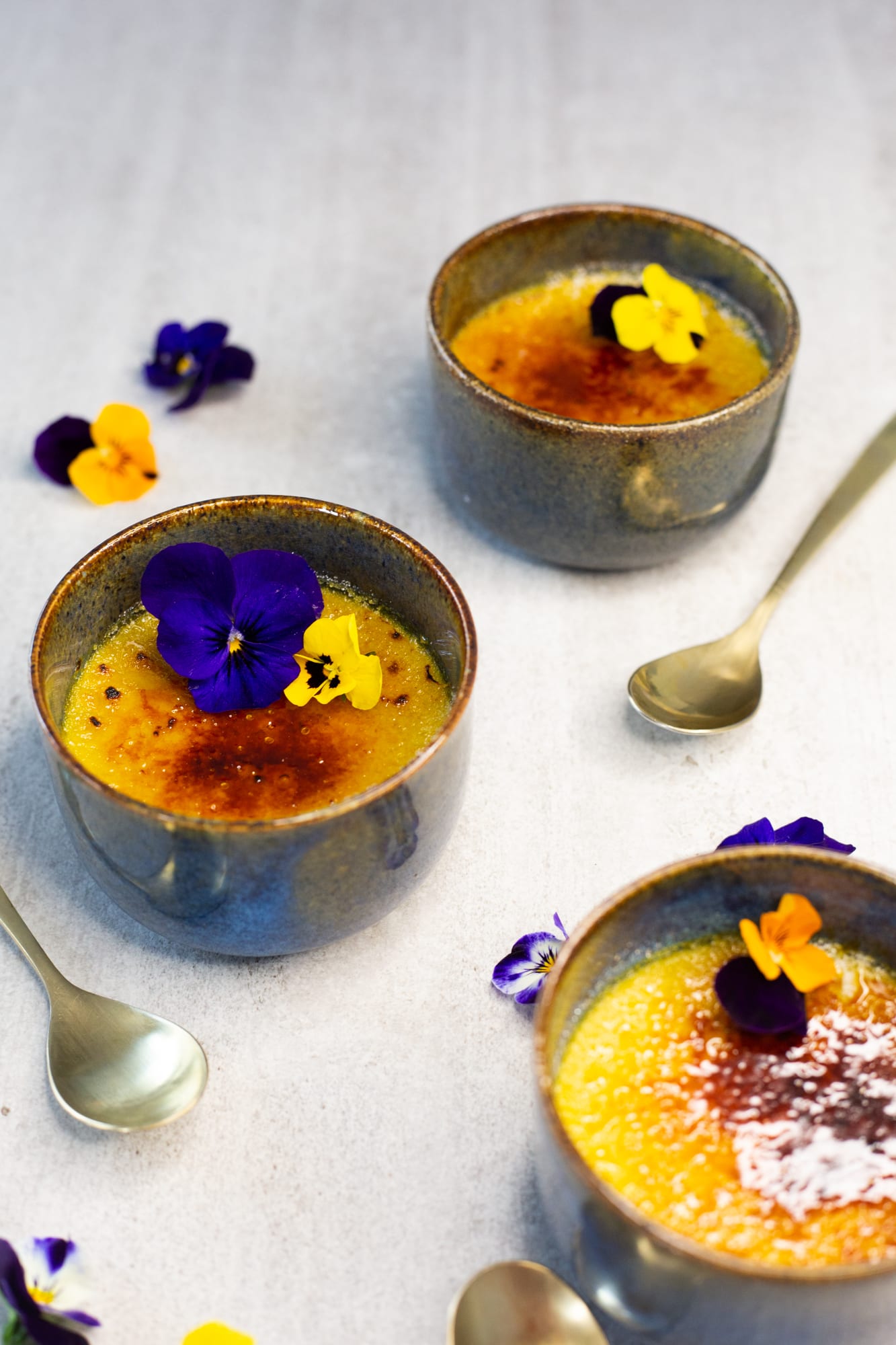 Three chai-spiced crème brûlées with violas in a diagonal row, from above at an angle