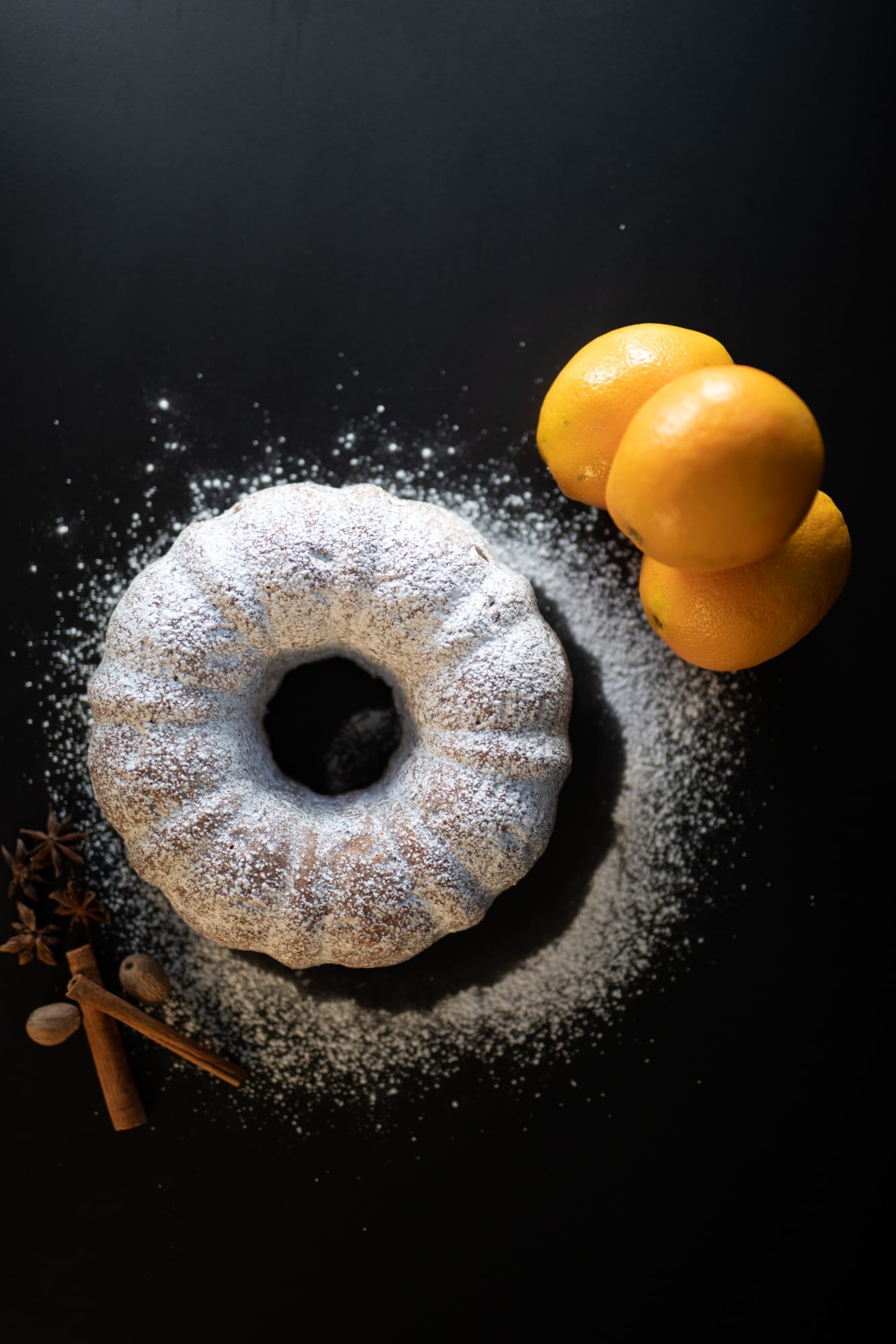 An orange spice pound cake dusted with icing sugar, with oranges, cinnamon and star anise, from above
