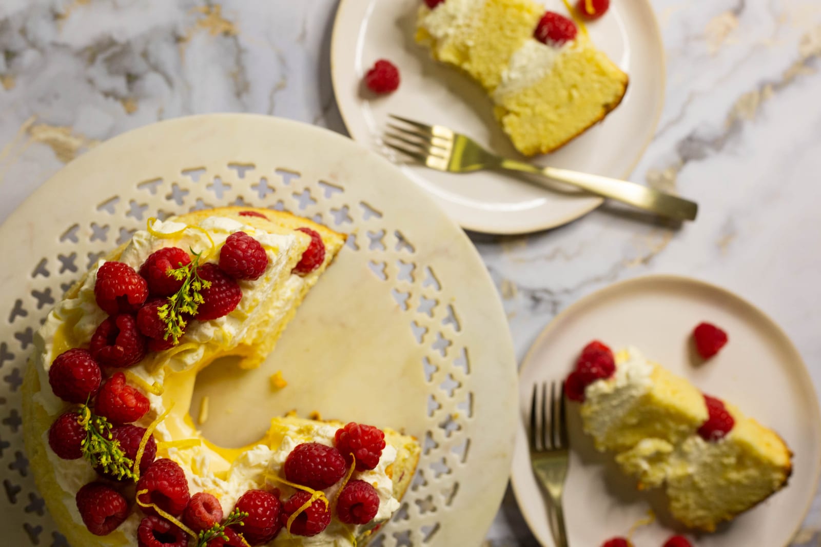 A cut lemon curd and raspberry chiffon cake on its stand with slices on plates and forks, from above