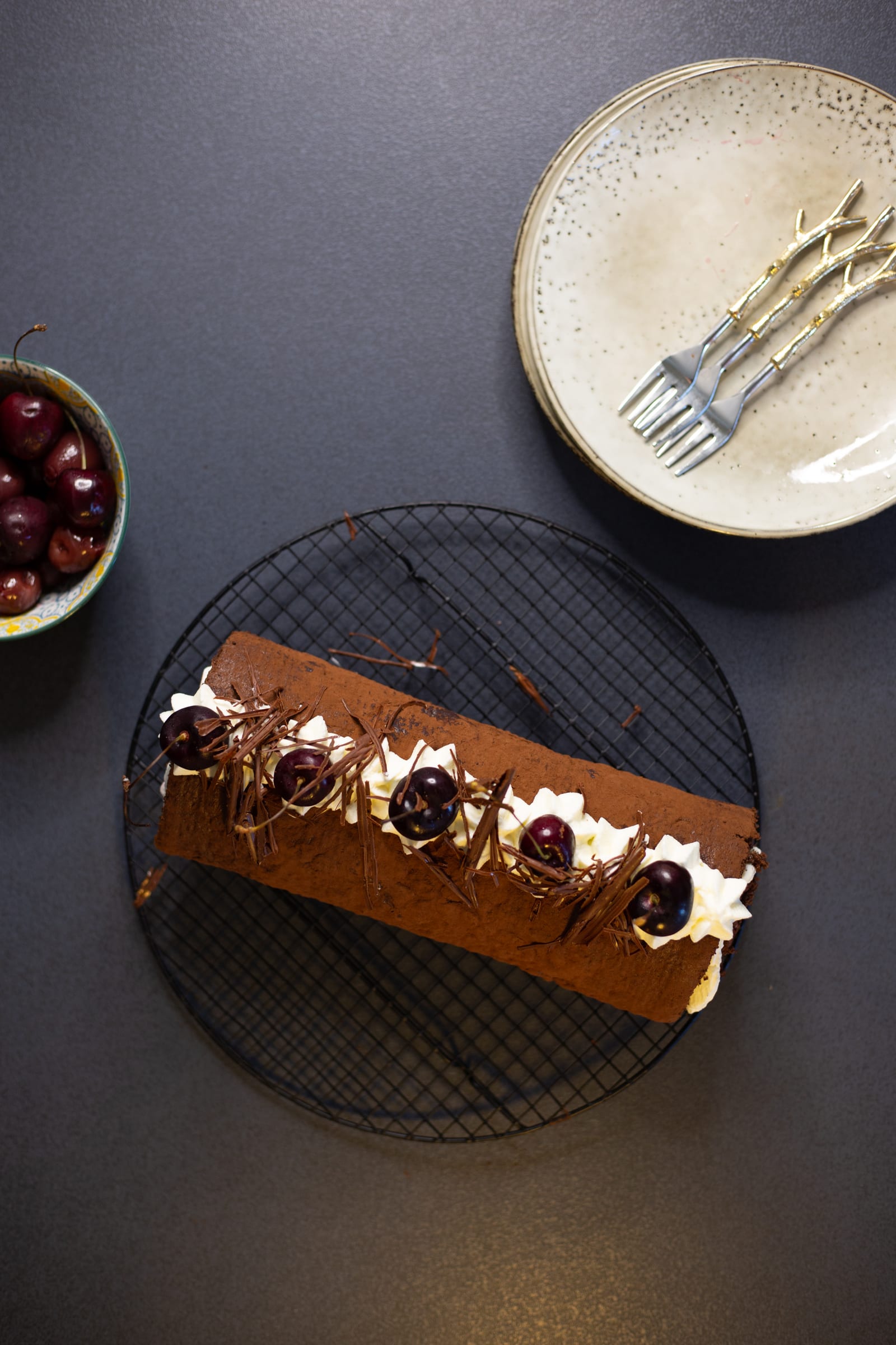 A decorated Black Forest Swiss roll on a round rack with plates and cherries, from above