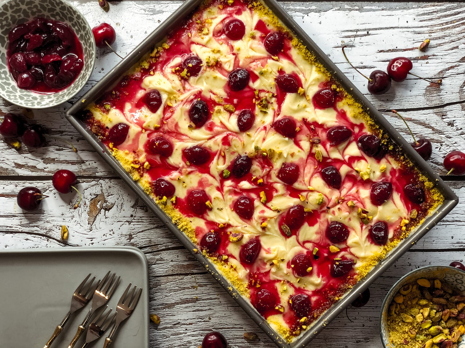 A pistachio and cherry sheet cake in its tin with bowls of cherries and pistachios and a plate of forks, from above