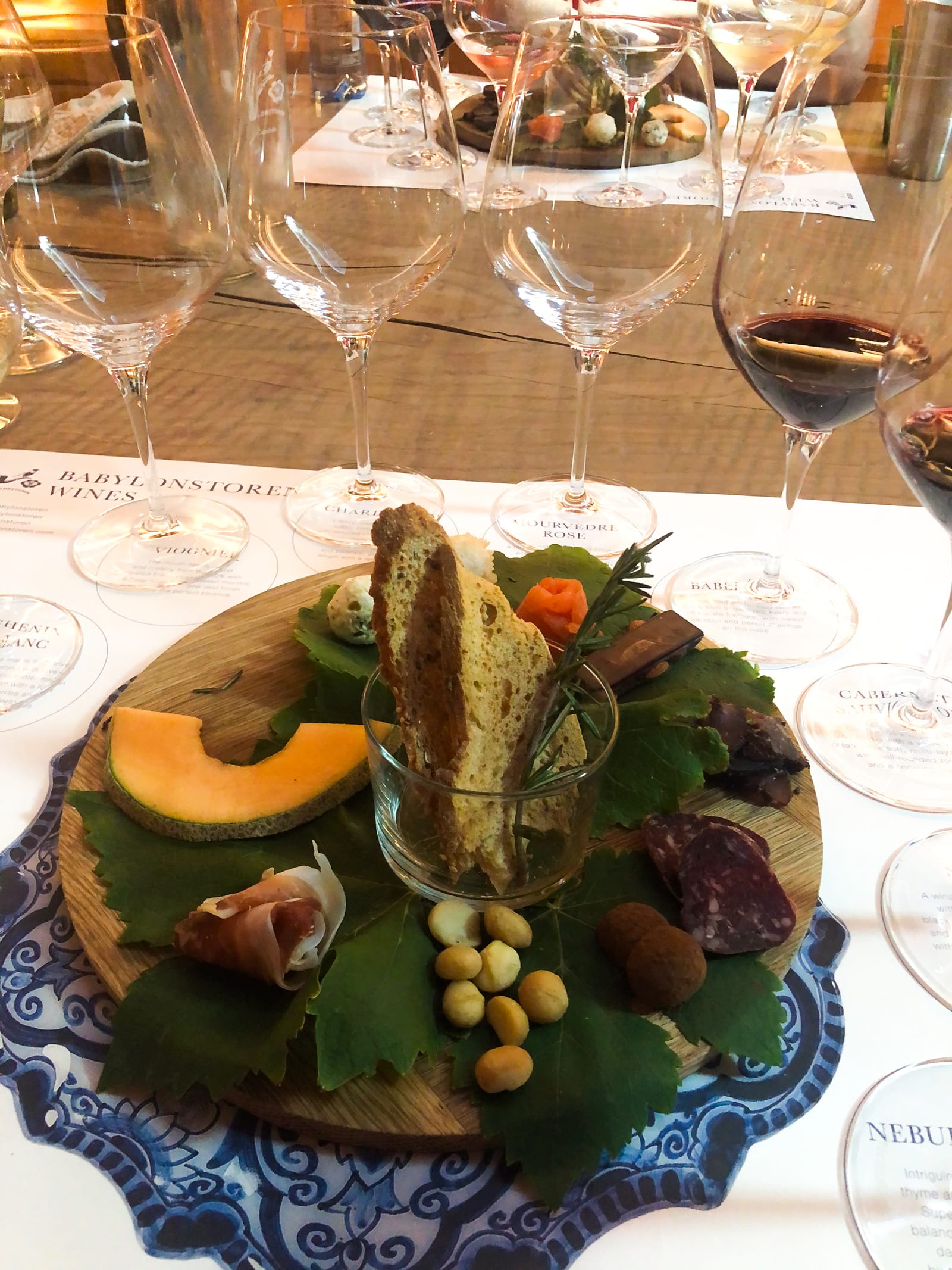 A wine-tasting pairing board of melon, prosciutto, salami, biltong, macadamias, chocolate and a rusk on vine leaves, ringed by Babylonstoren wine glasses