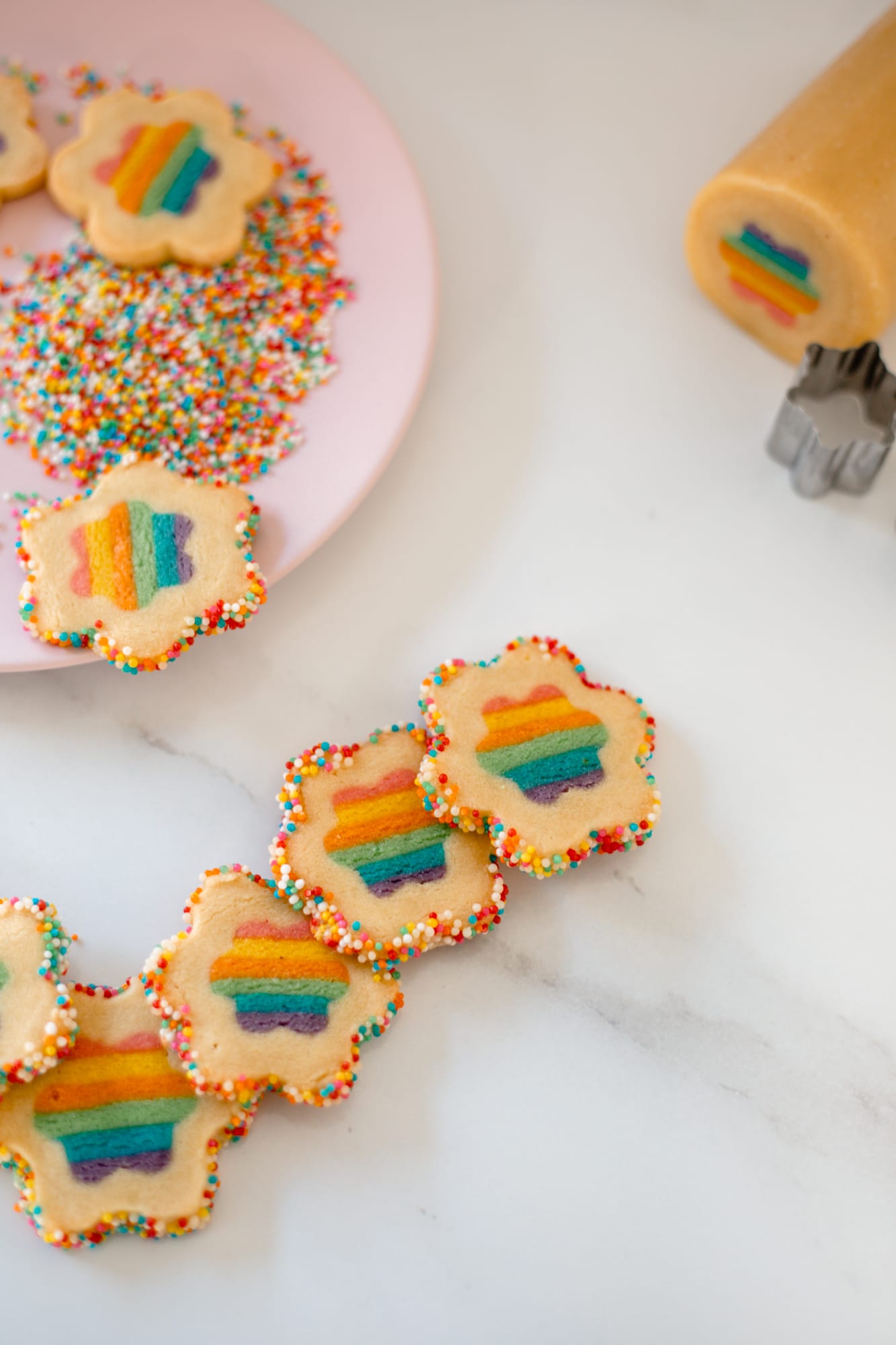 Rainbow cookies beside the log of dough with its rainbow centre and a flower cutter