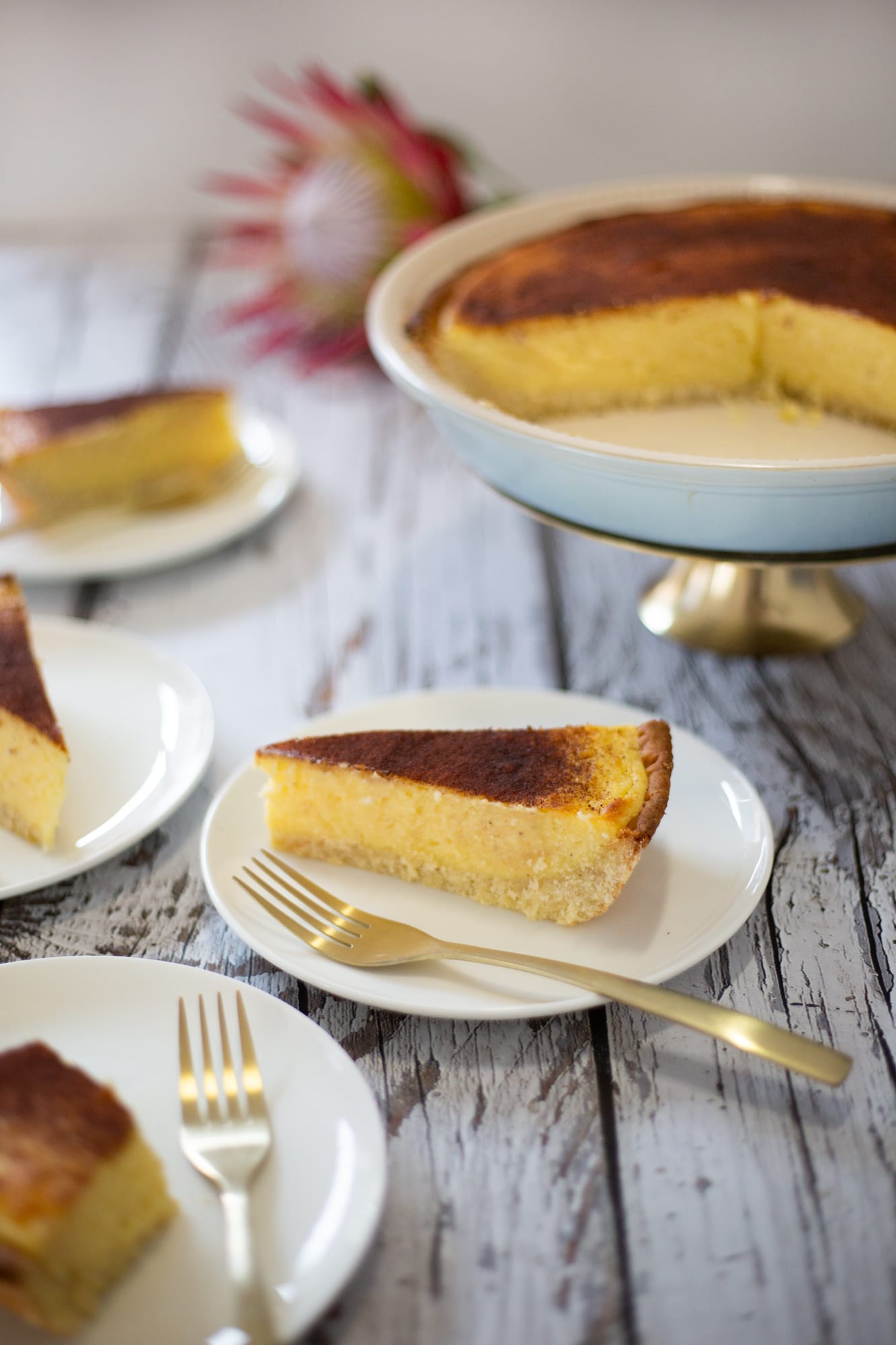 A cut melktert on a cake stand with slices on white plates and gold forks