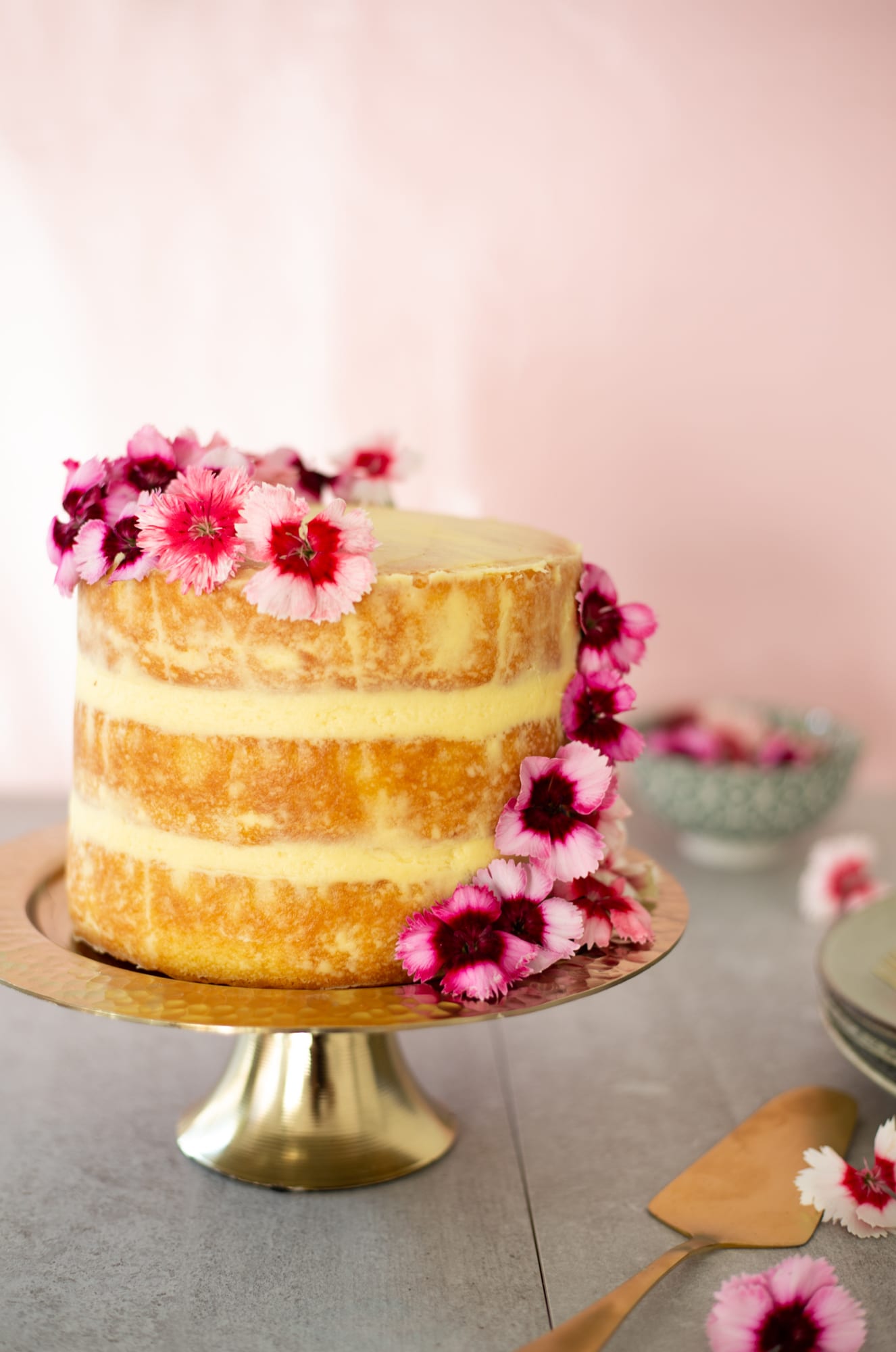 A three-layer lemon curd cake with pink flowers on a gold stand against a pink wall