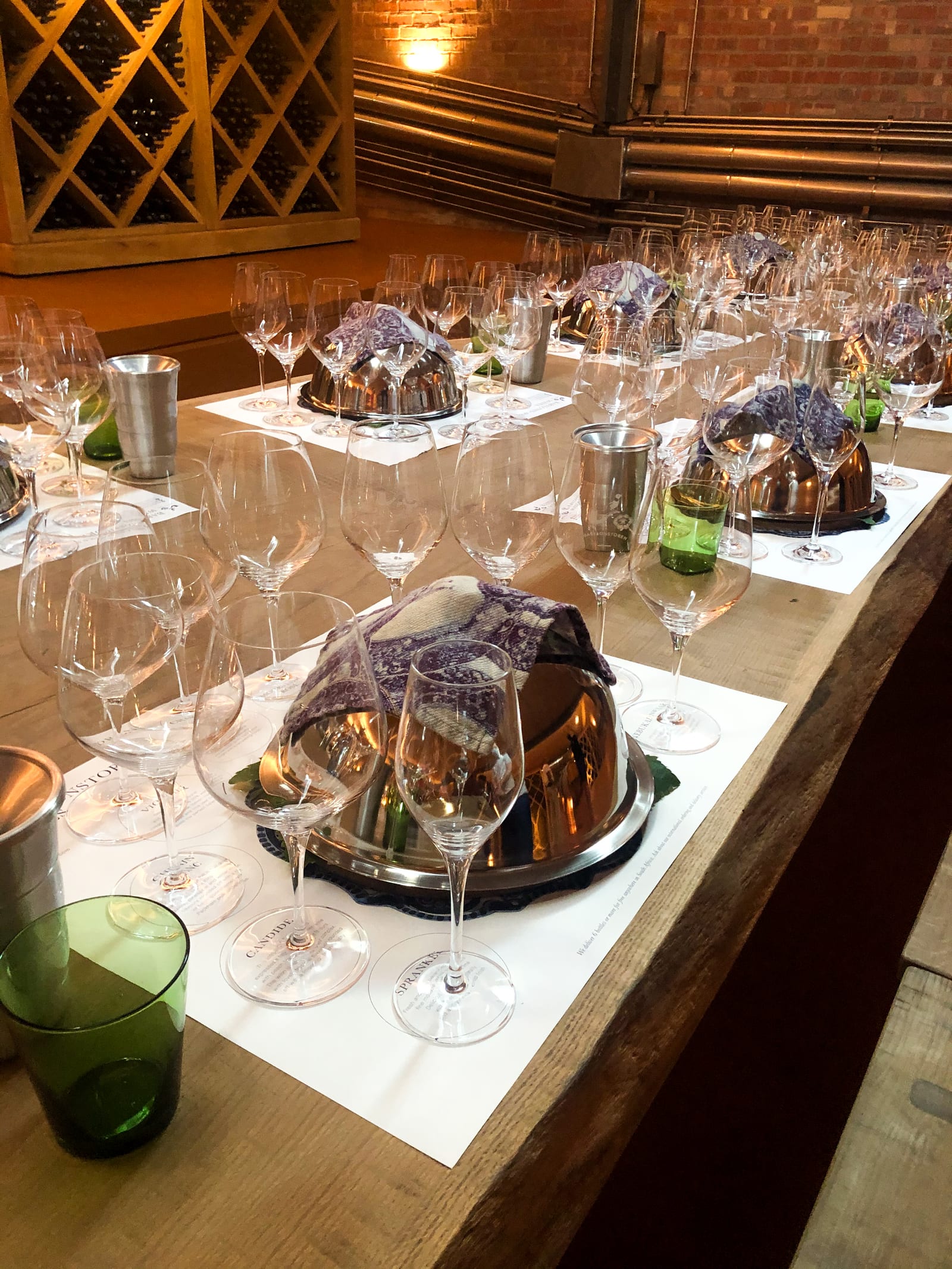 A long wooden tasting table in the brick-walled wine cellar, set with rows of wine glasses and covered food-pairing platters