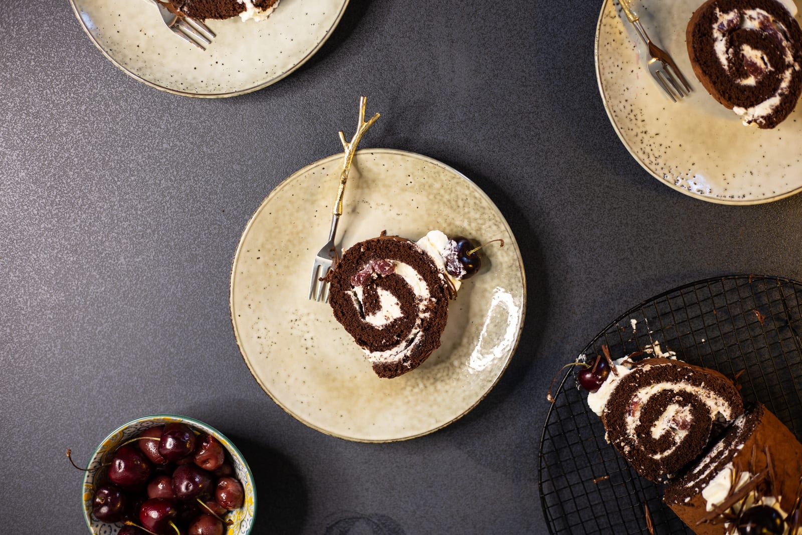Spiral slices of Black Forest Swiss roll on cream plates with gold forks and cherries, from above