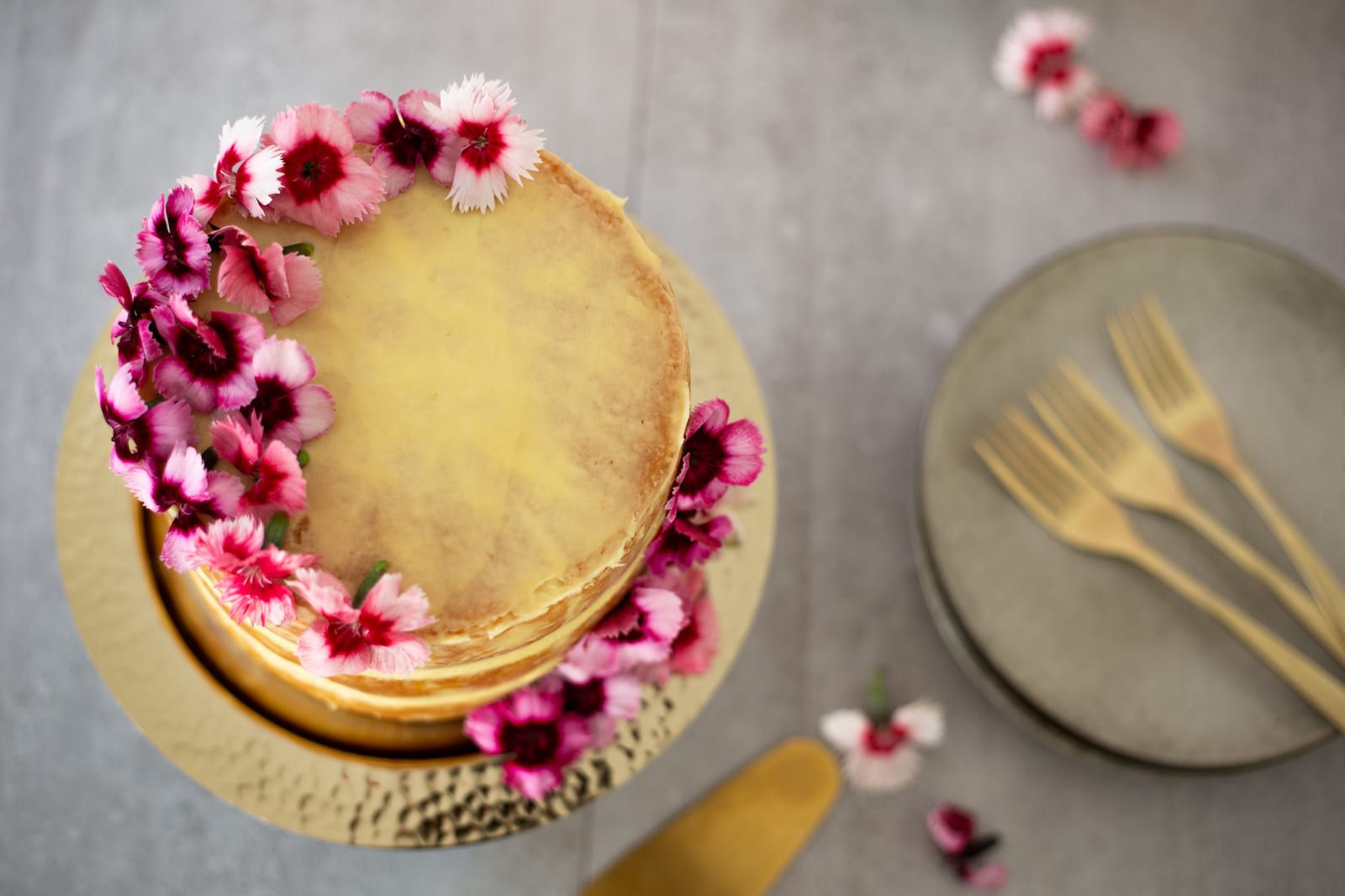 A lemon curd cake with flowers beside plates and gold forks, from above