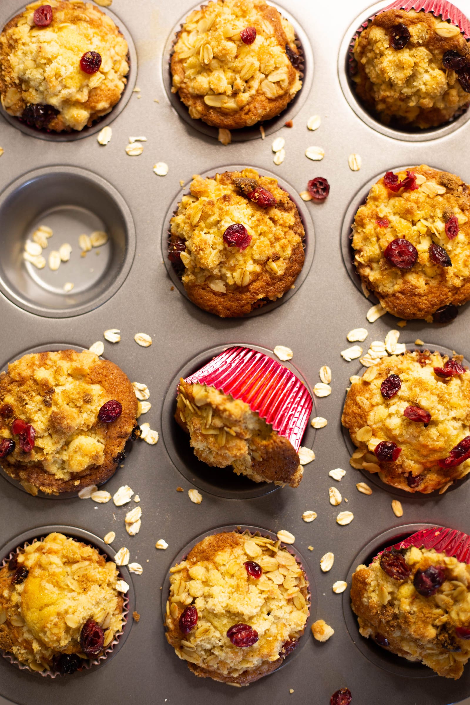 Close-up of a tin of cranberry crumble muffins, one tipped to show its red case, from above