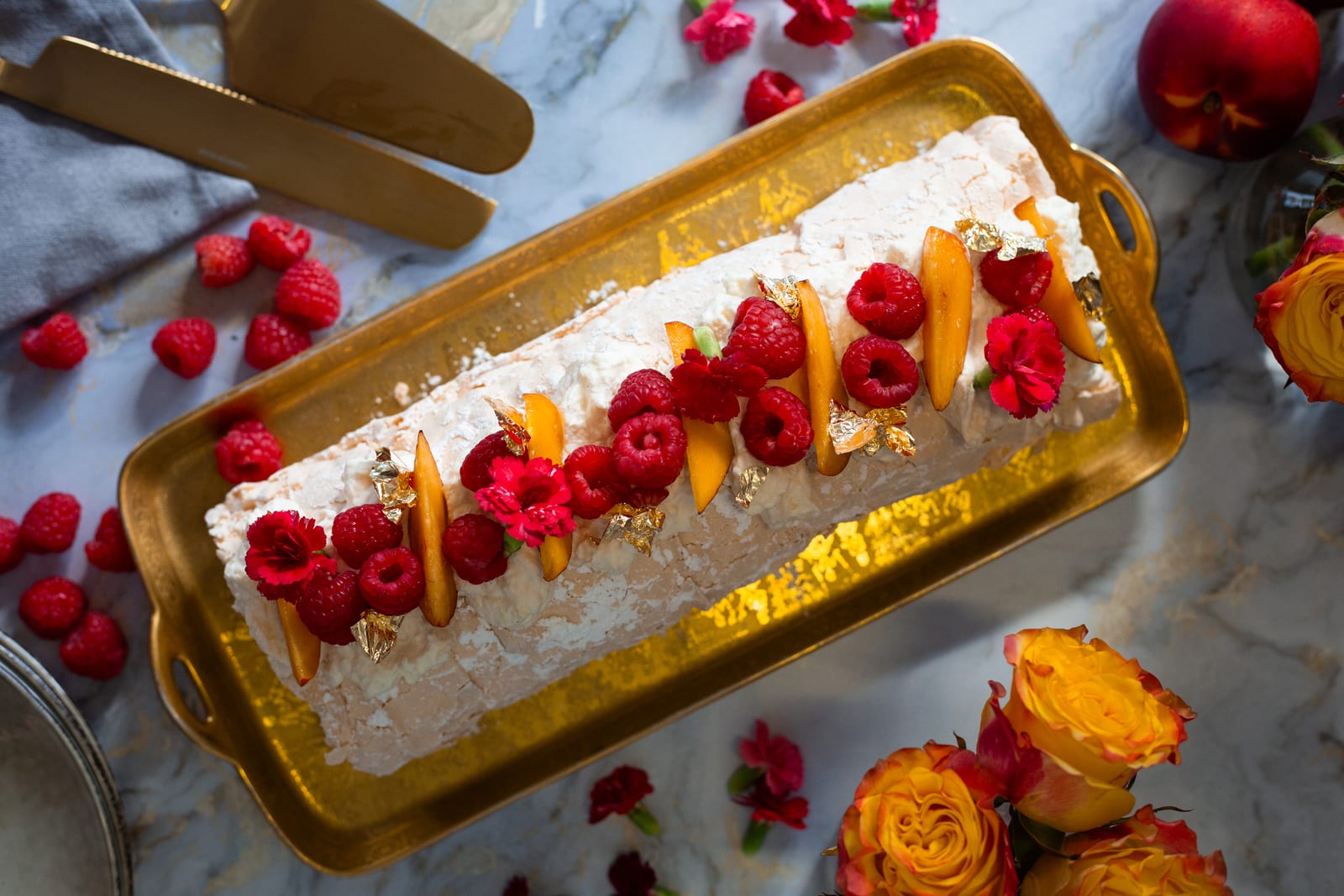 Raspberry and nectarine pavlova roulade from above on a gold tray, topped with raspberries, nectarine slices and gold leaf