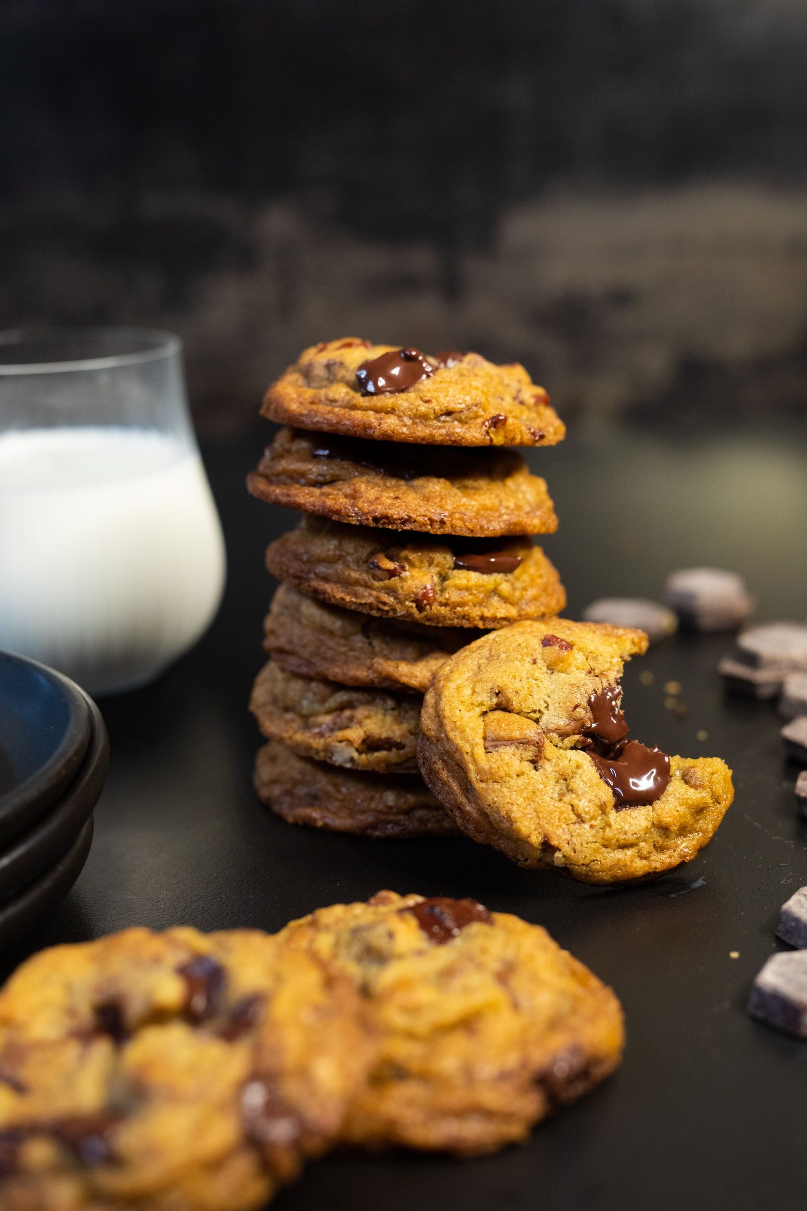 A tall stack of chocolate chip cookies with a bitten cookie leaning on it, milk and chocolate chunks beside it, from the side