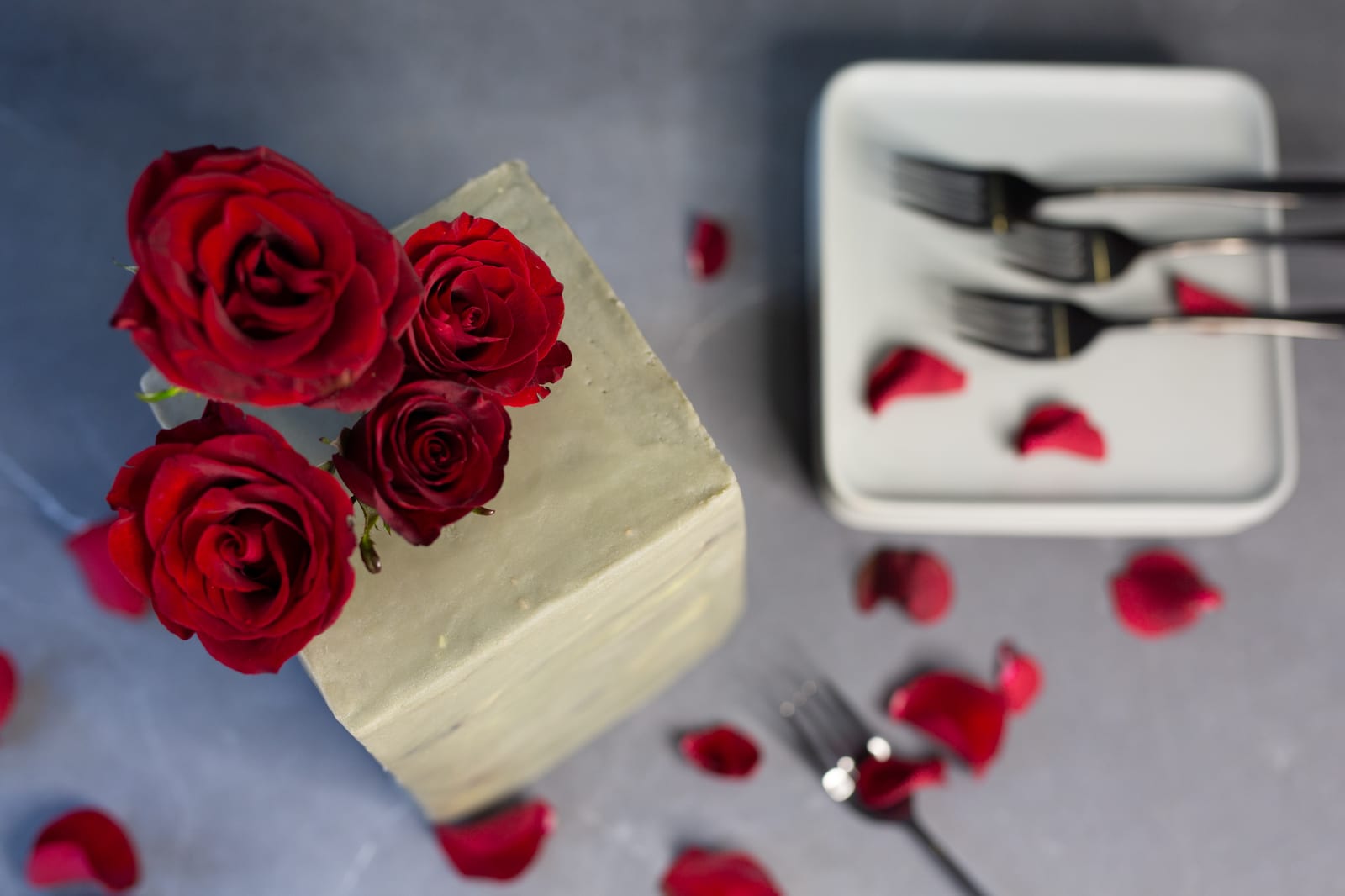 A tall decadent red velvet cake in white frosting topped with red roses, petals and forks around it