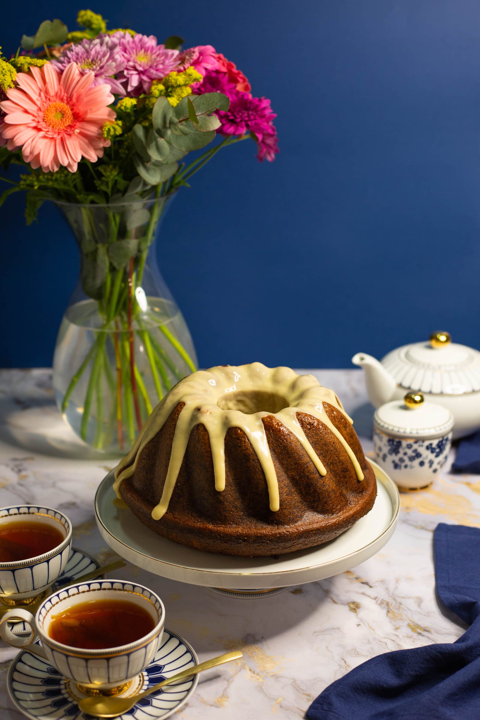 A glazed rooibos and honey bundt on a white cake stand beside a teapot, cups of tea and pink gerberas