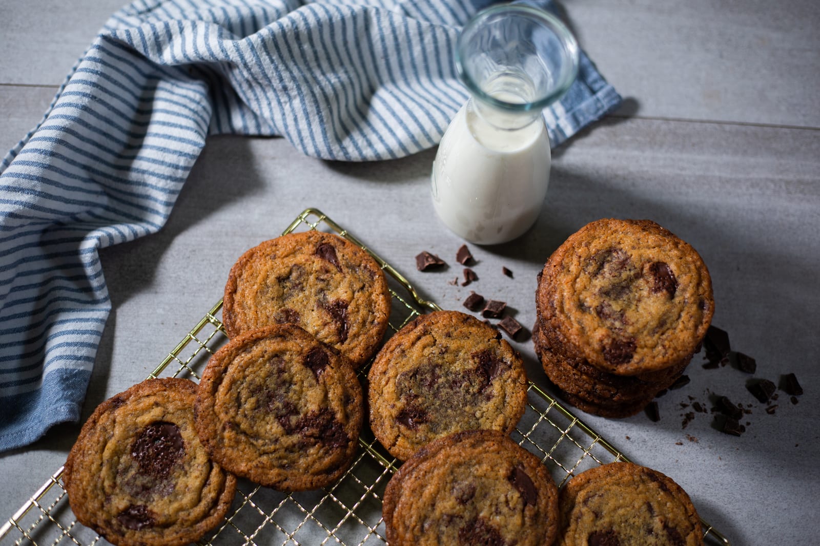 Pan banging chocolate chip cookies on a gold rack beside a carafe of milk and a striped towel