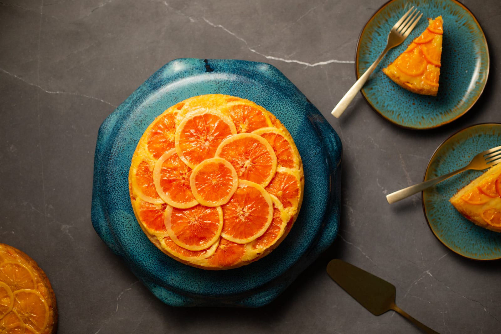 A whole orange upside down cake on a teal plate beside small plates of slices and forks, from above