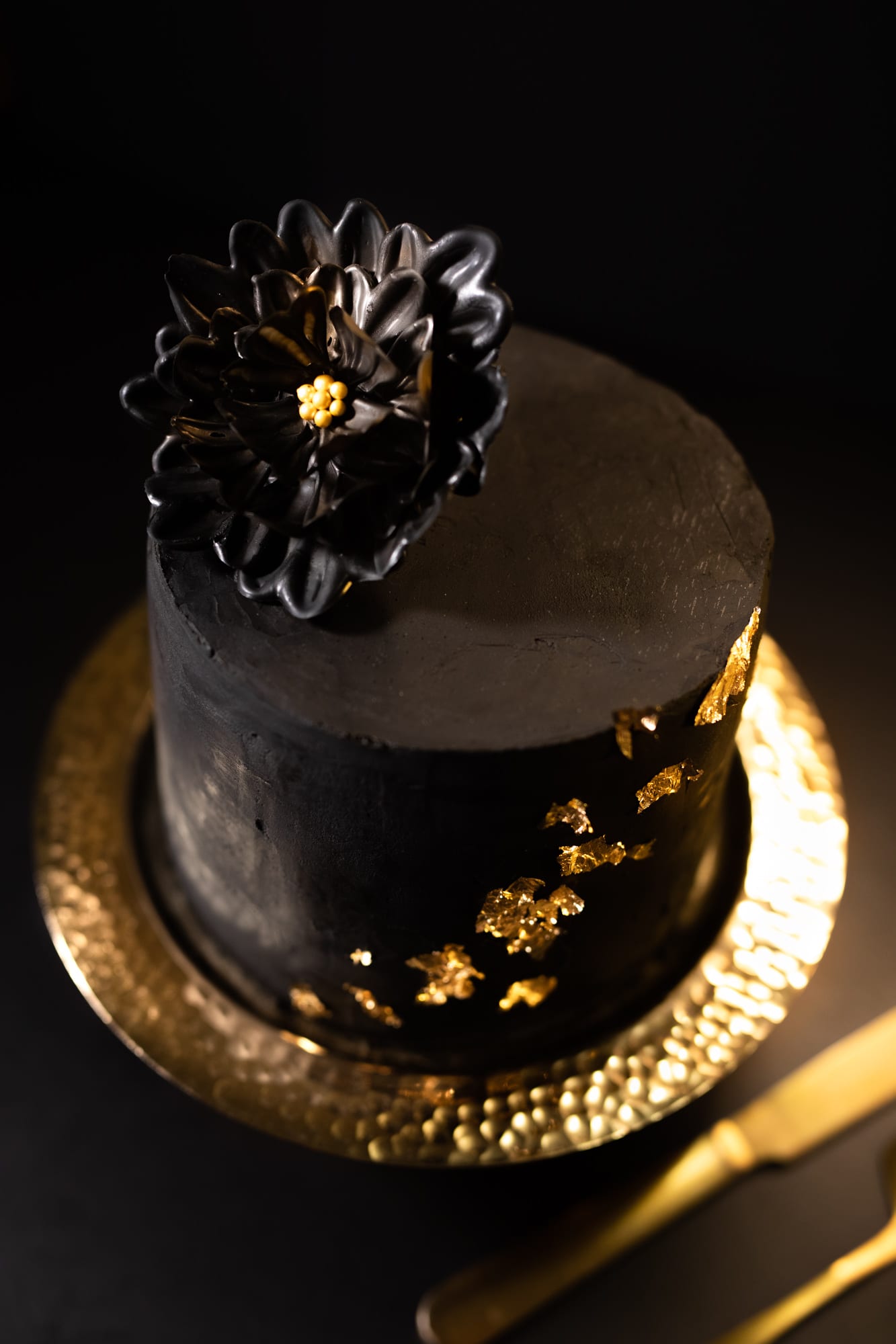 The black chocolate flower and gold leaf on top of a black velvet cake on a hammered gold stand