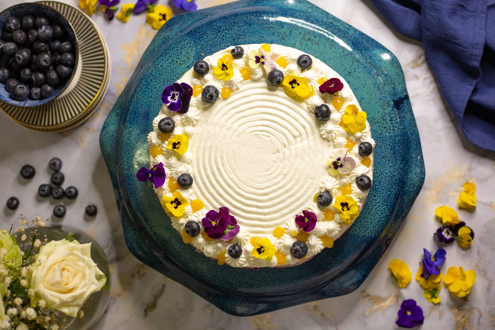 A lemon and blueberry cheesecake from above, ringed with edible flowers and blueberries on a teal stand