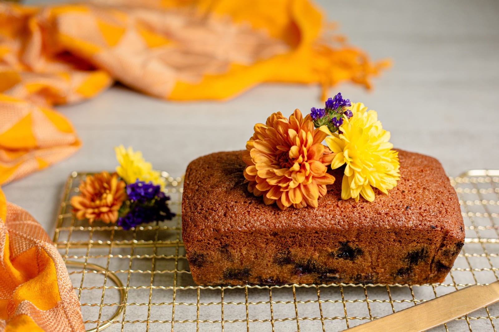 A lemon and blueberry loaf with flowers on a gold rack over orange silk