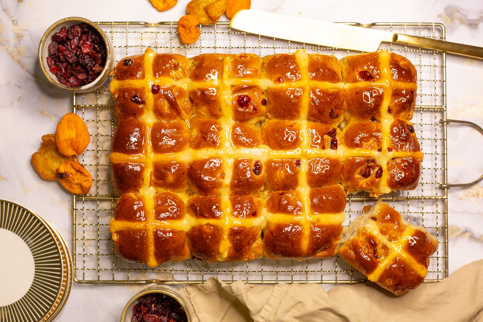 A tray of Turkish apricot and cranberry hot cross buns on a wire rack, with one bun pulled away