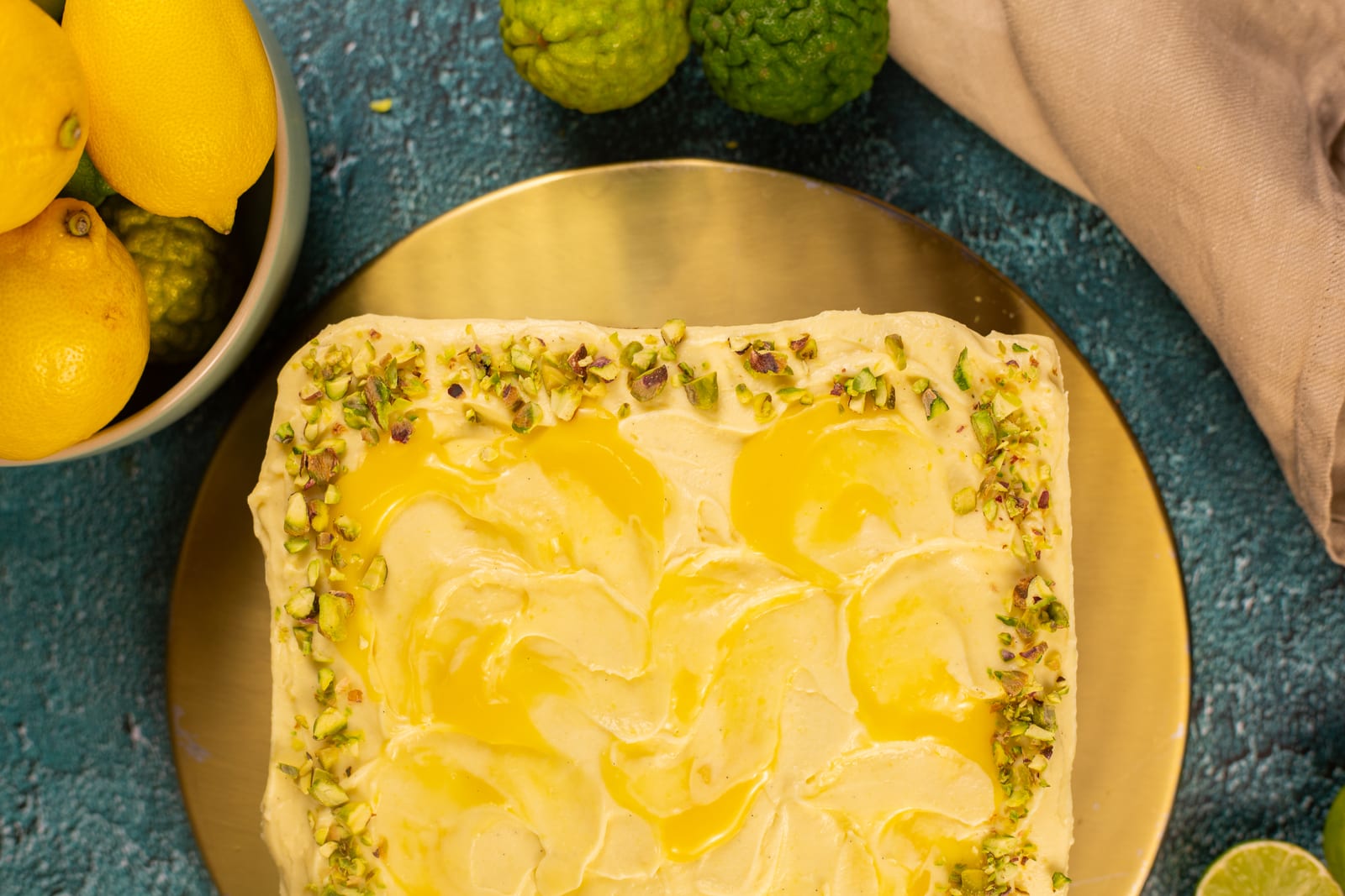 A pistachio and lemon traybake from above on a gold board, beside a bowl of lemons and green citrus