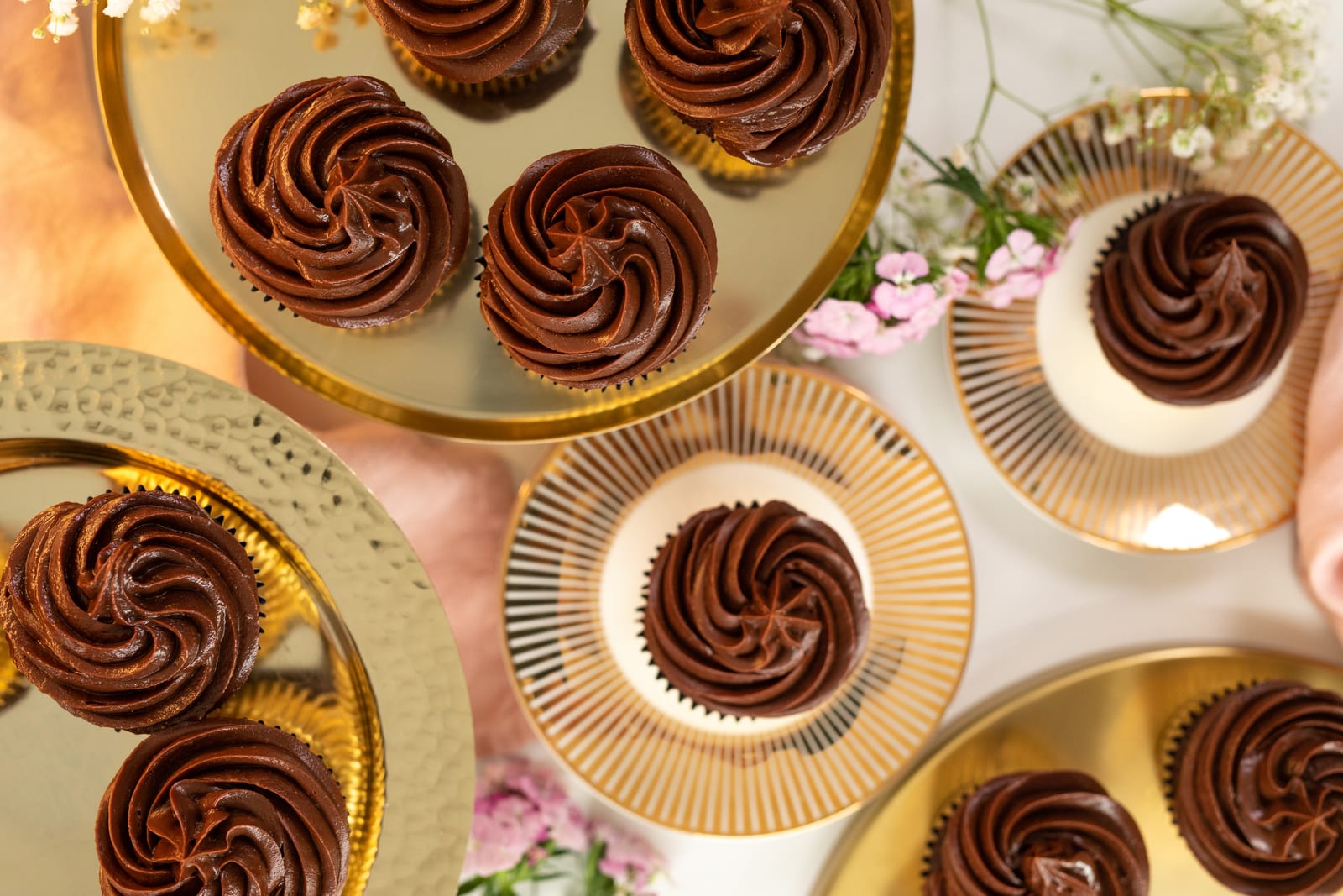Swirls of chocolate ganache on ultimate triple chocolate cupcakes on gold and white plates with pink flowers, from above