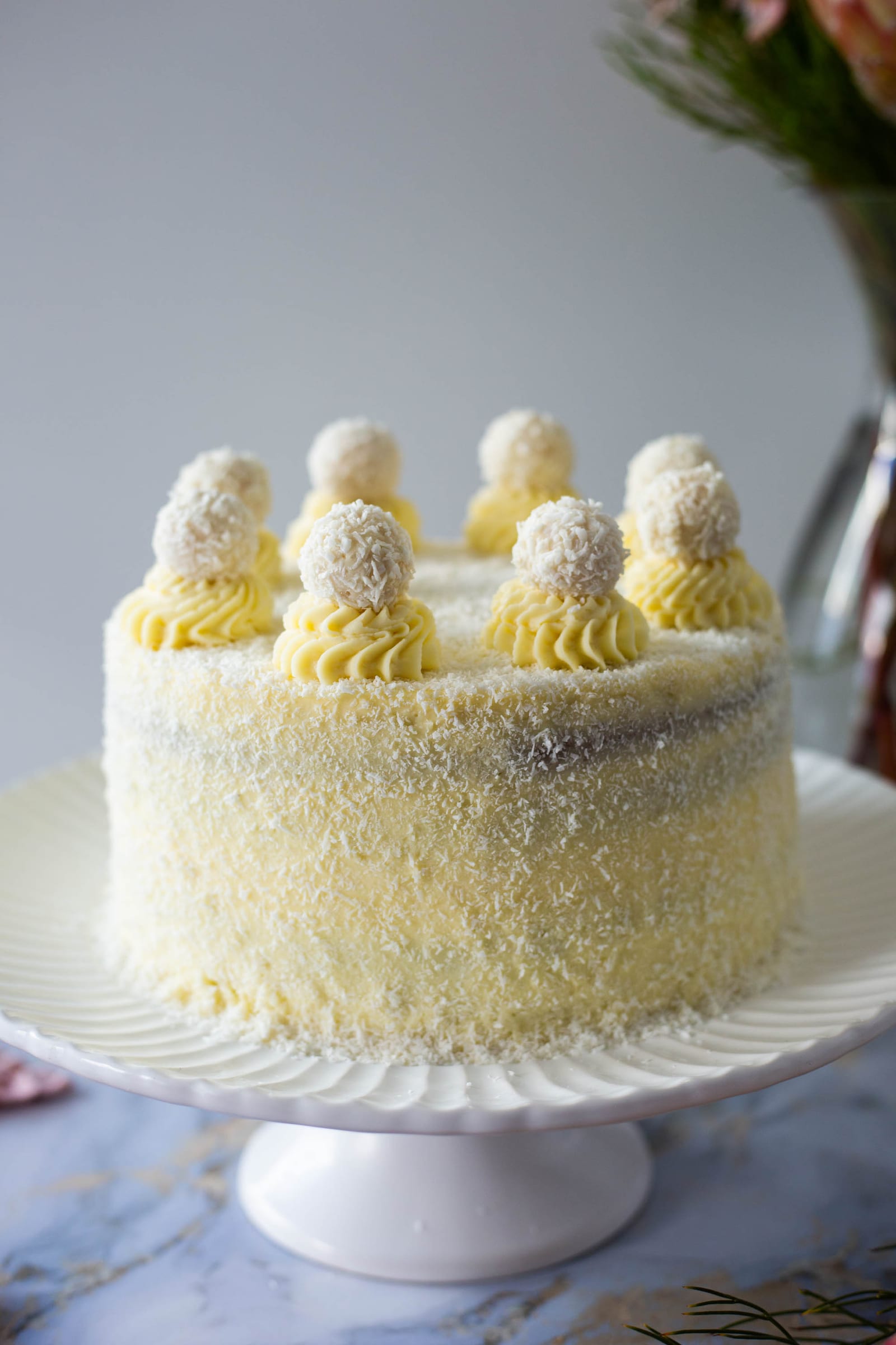 A coconut-covered Raffaello cake on a white cake stand, from the side