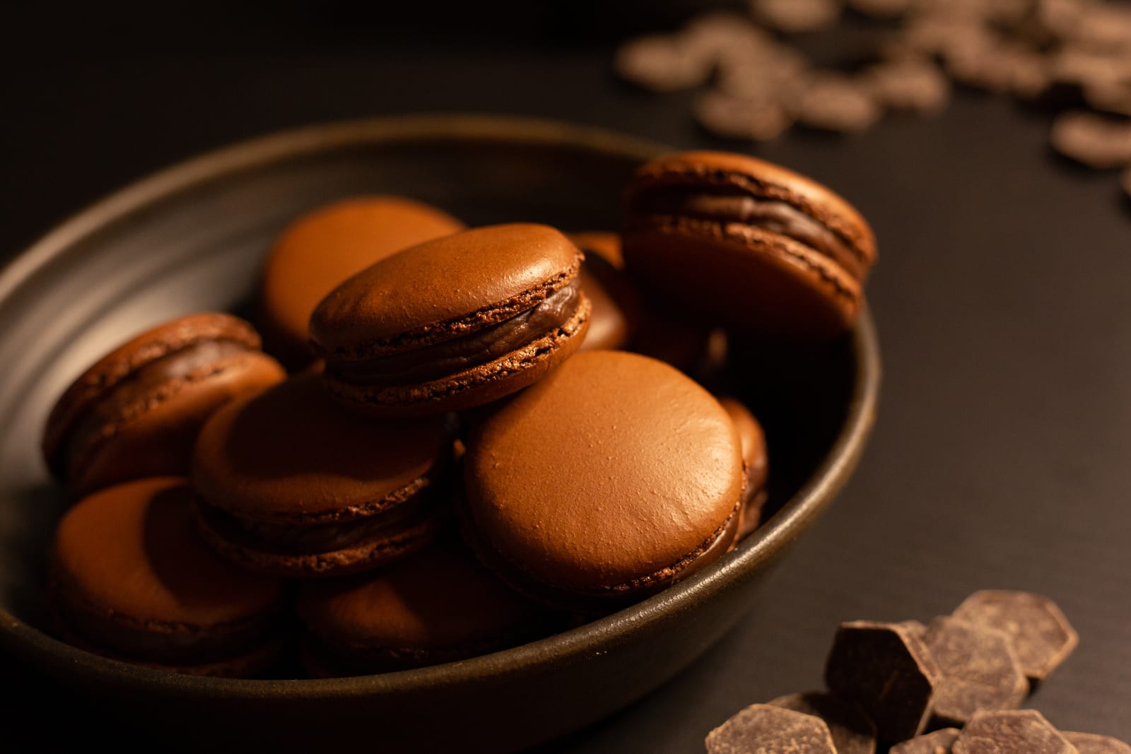 A bowl piled with chocolate macarons beside scattered chocolate pieces