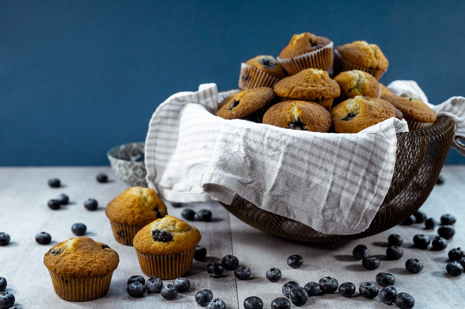 A basket of one bowl blueberry muffins with blueberries scattered in front, against a teal backdrop