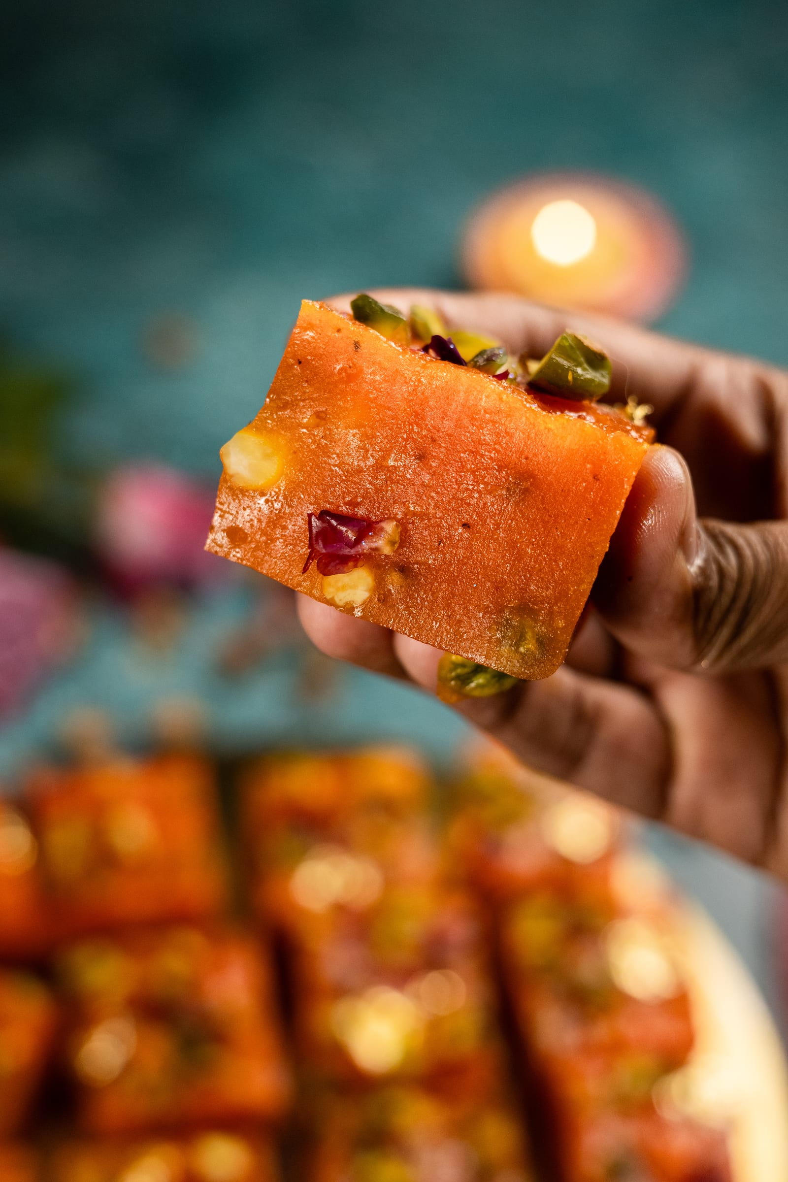 A hand holding up a block of halwa, showing the nuts set inside, with a lit diya behind