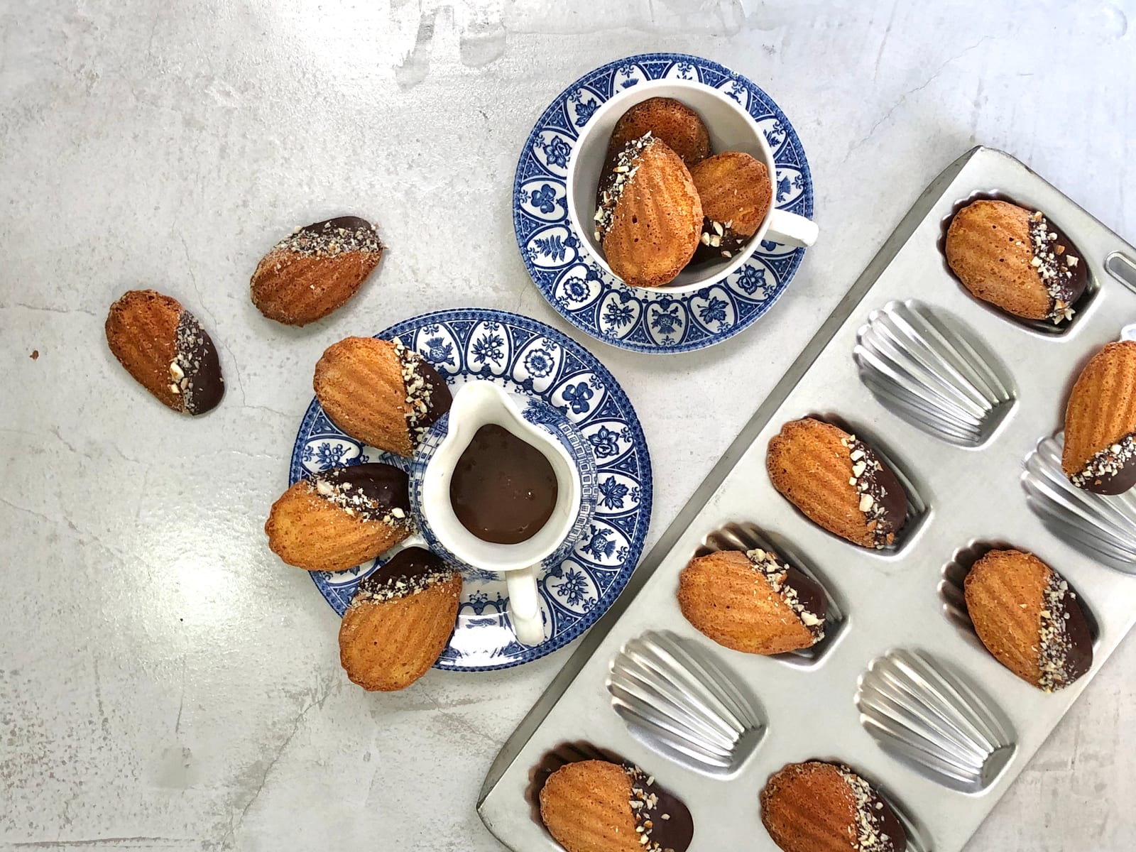 Brown butter and almond madeleines dipped in chocolate beside a madeleine tin, a jug of chocolate and a cup and saucer, from above
